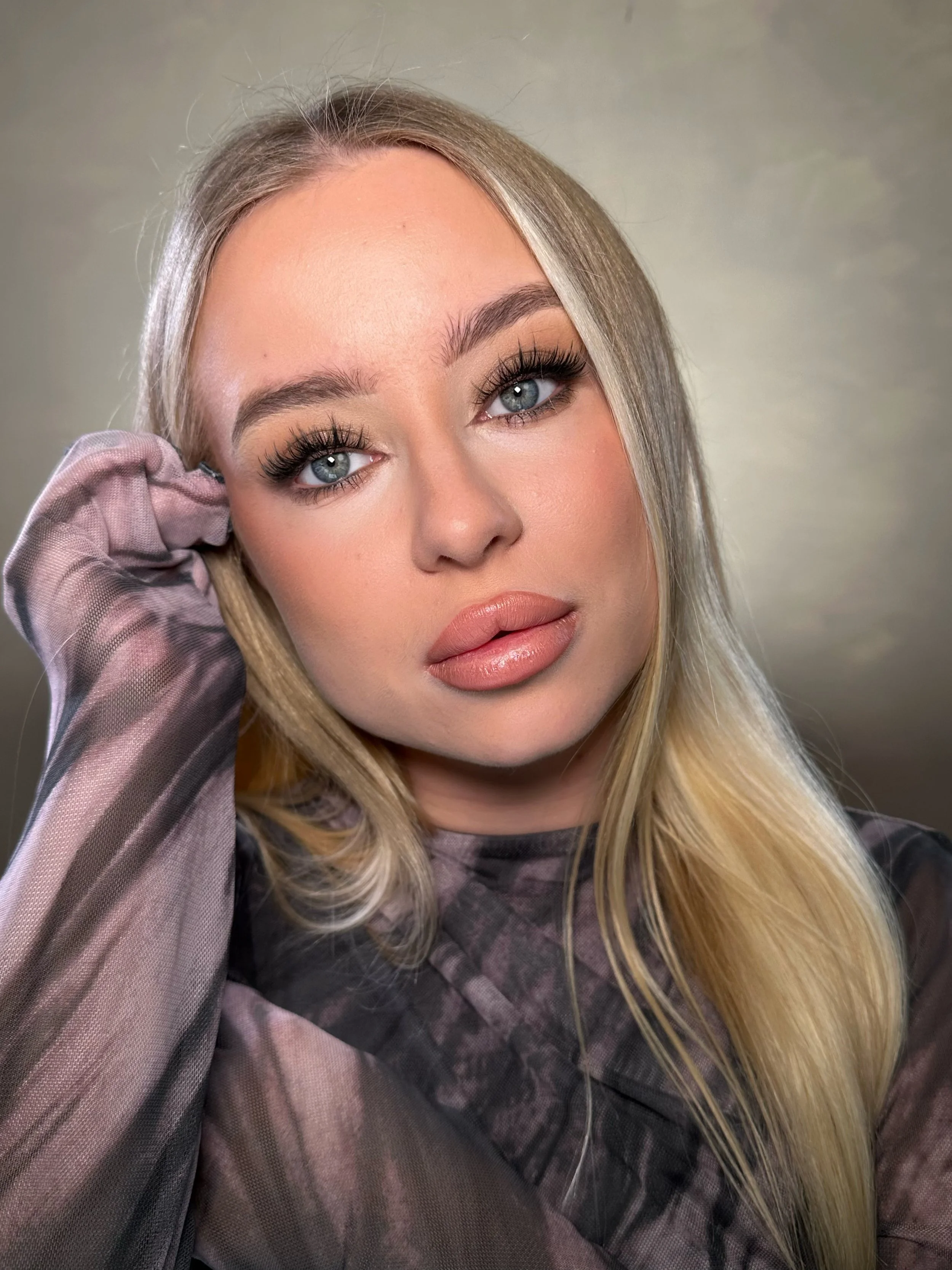 Close-up of a person with long blonde hair, wearing a patterned shirt, posing with one hand on their head, looking thoughtfully.