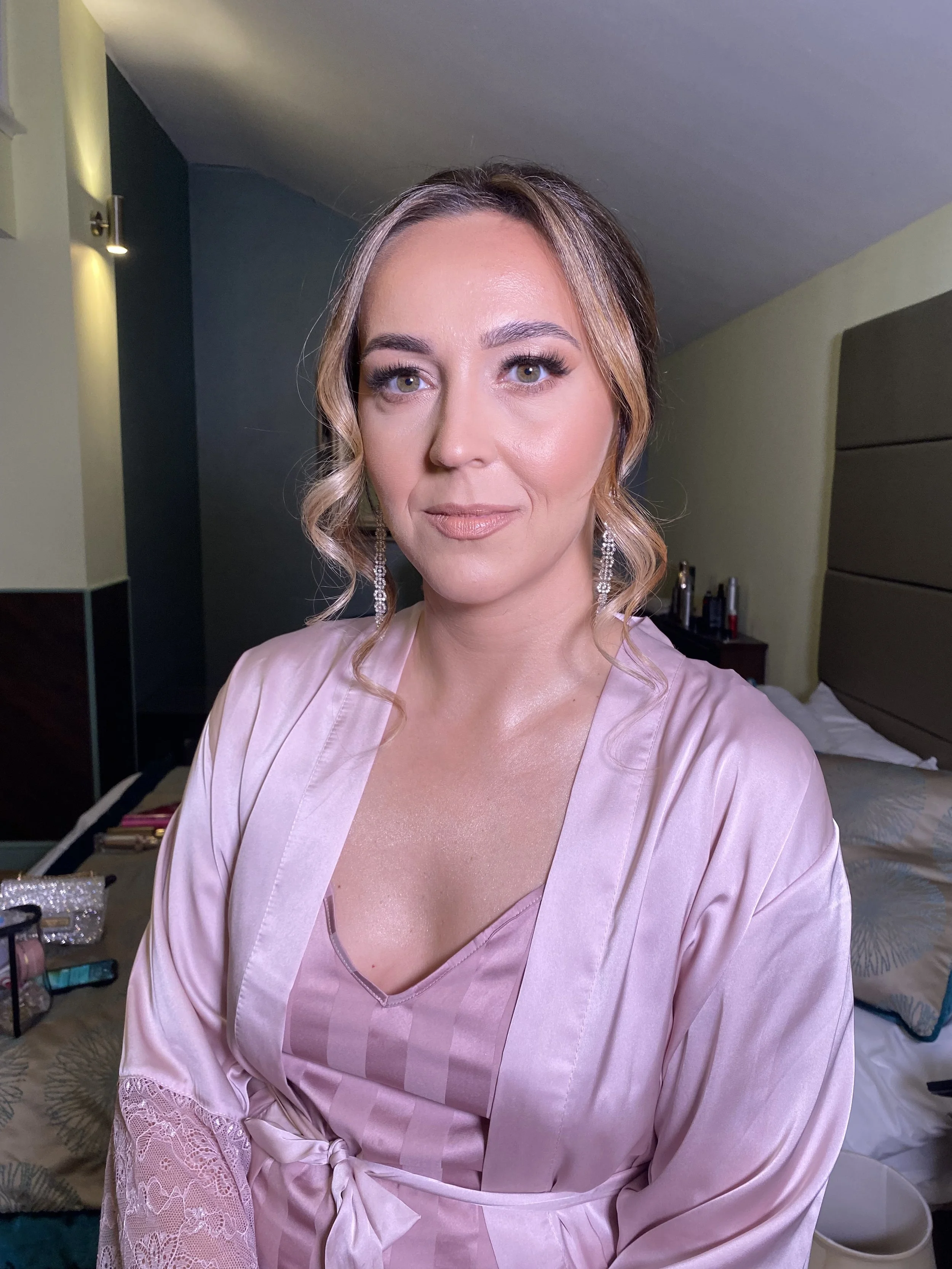 A woman with styled hair and makeup, wearing a pink satin robe with lace detailing, sitting on a bed in a hotel room with makeup and personal items on a side table.