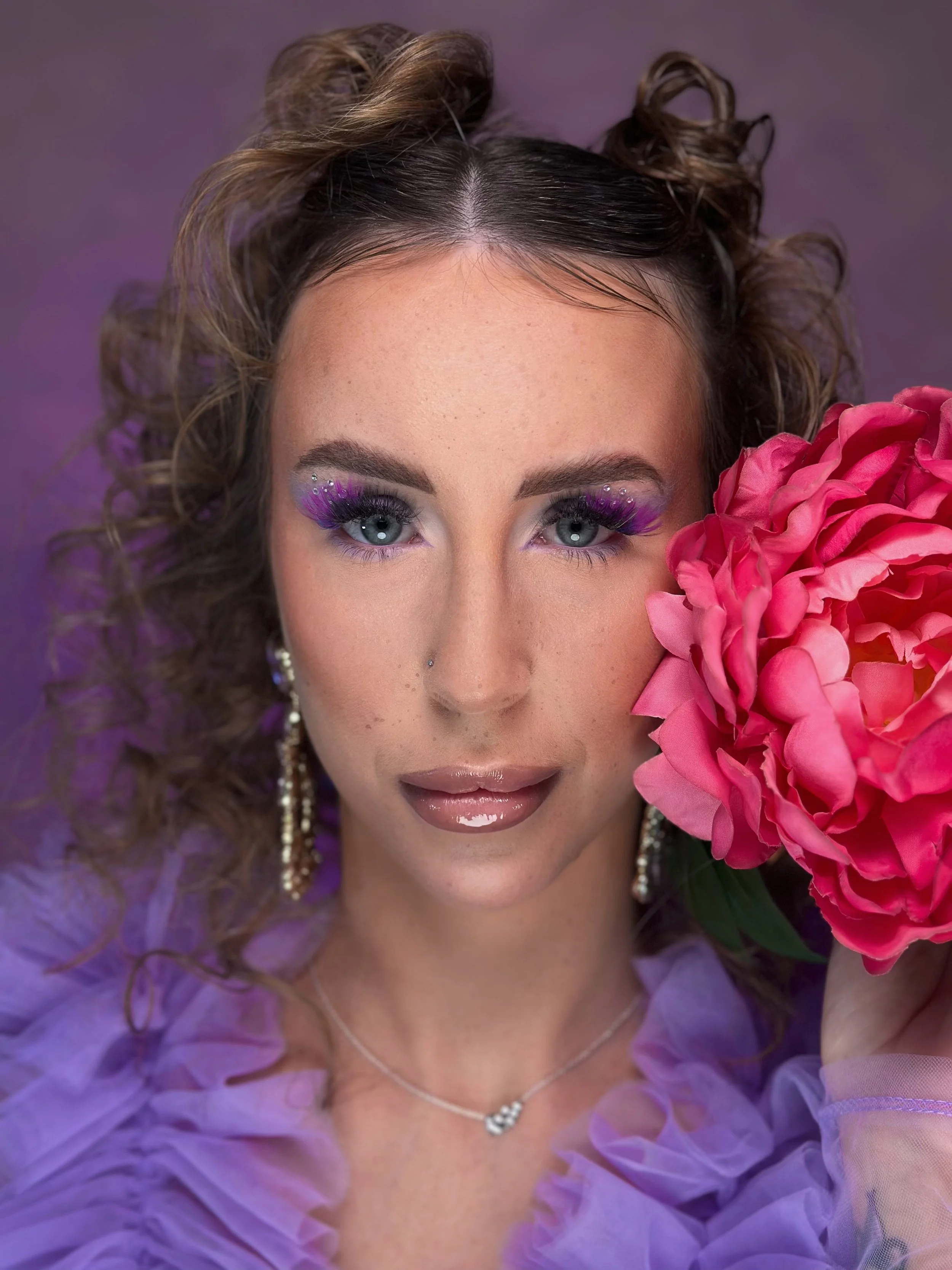 A woman with curly hair styled in buns, wearing vibrant purple eye makeup and glossy lips, holding a large pink flower. She is dressed in a sheer purple top and accessorized with statement earrings and a delicate necklace.