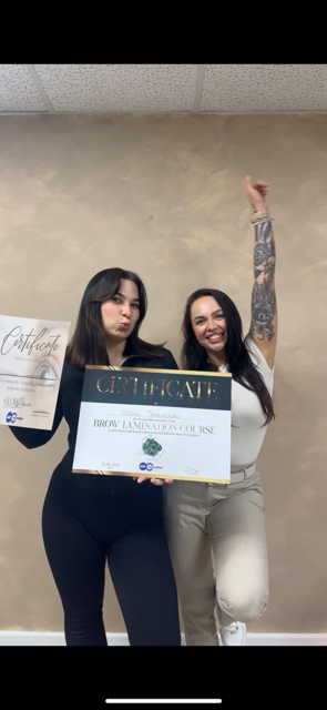 Two women standing indoors celebrating while holding certificates for a brow lamination course.