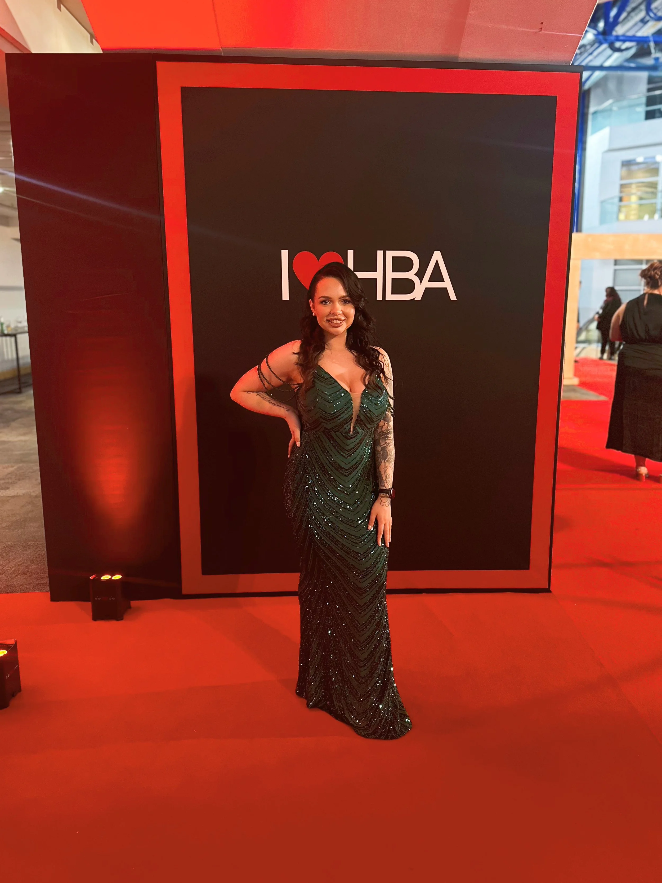 Woman in green dress at red carpet event in front of 'I Heart HBA' sign