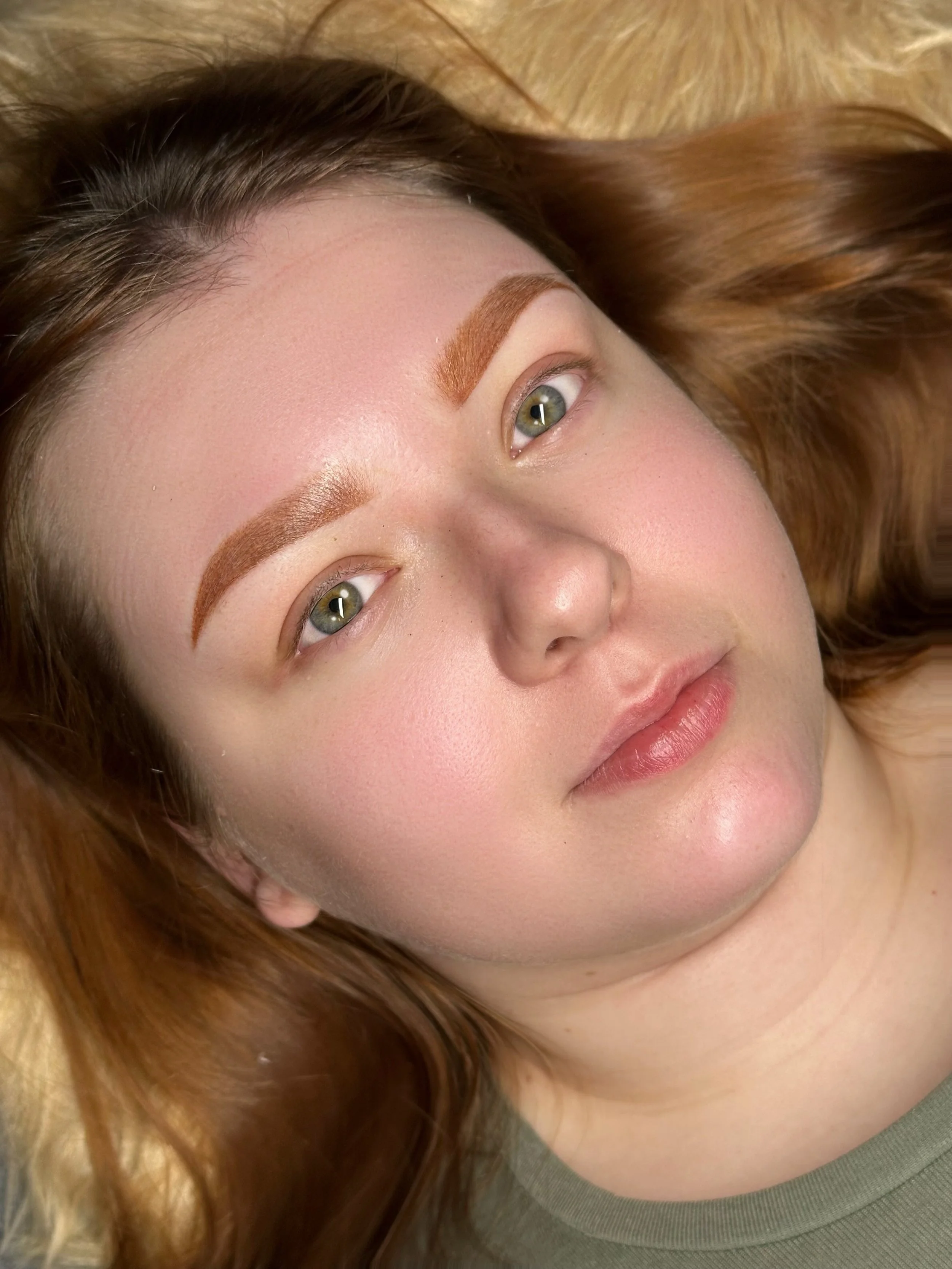 A woman with light skin and red hair lying down, looking upwards. She has well-defined eyebrows and is wearing a green shirt. Her expression is neutral and her hair is spread around her head.