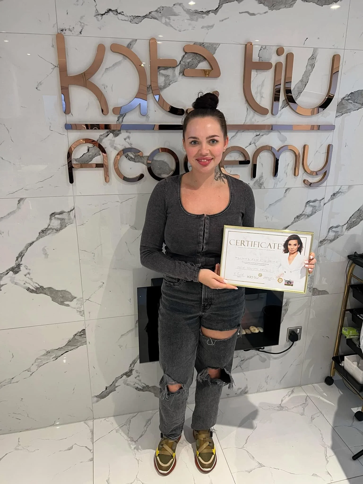 Person holding a certificate in front of "Kate Liu Academy" sign, wearing casual clothing, standing on marbled floor.