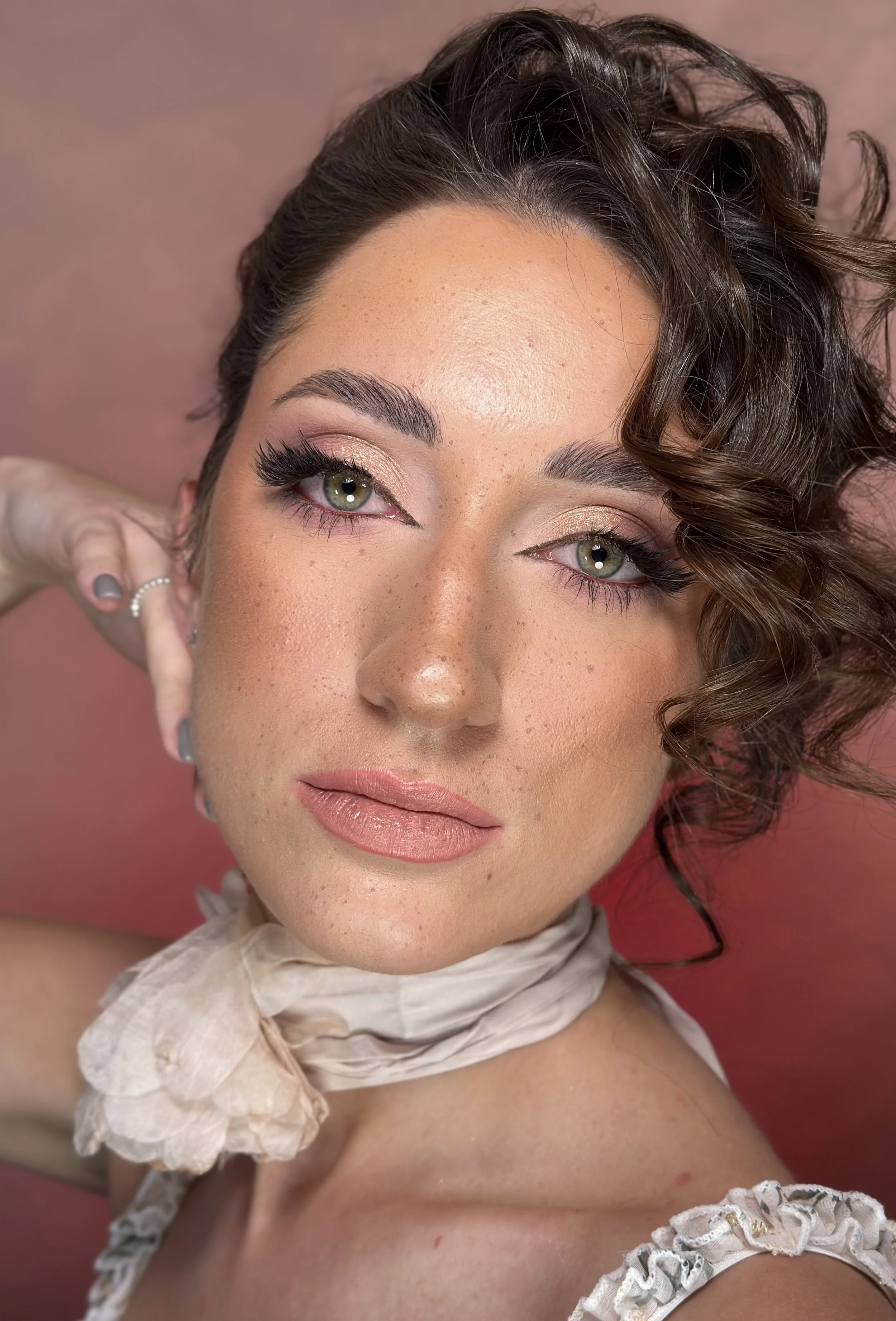 Close-up of a woman with curly brown hair, green eyes, and makeup, posing against a pink background.