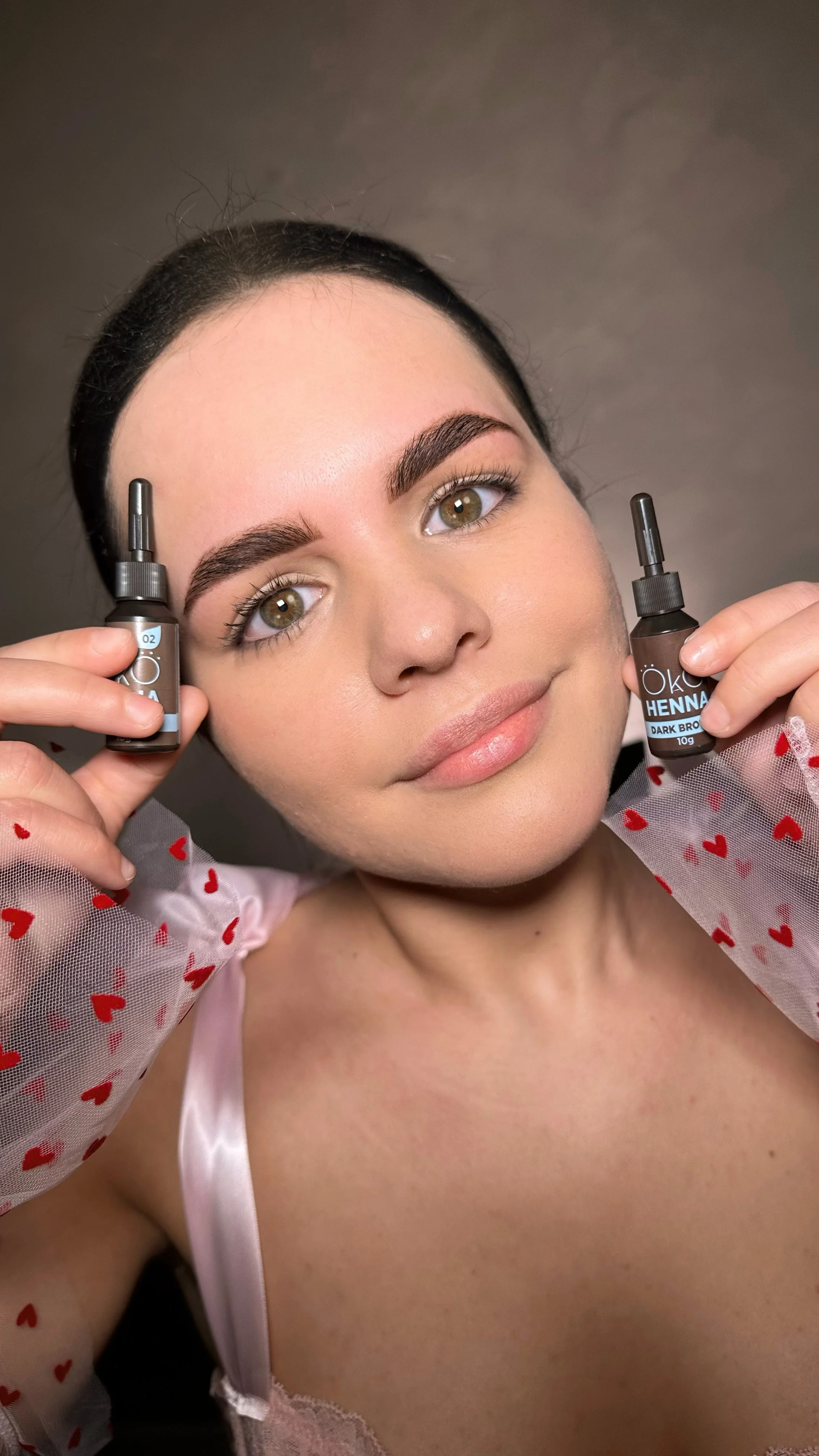 Person holding two bottles of eyebrow dye, wearing a pink top with red hearts.