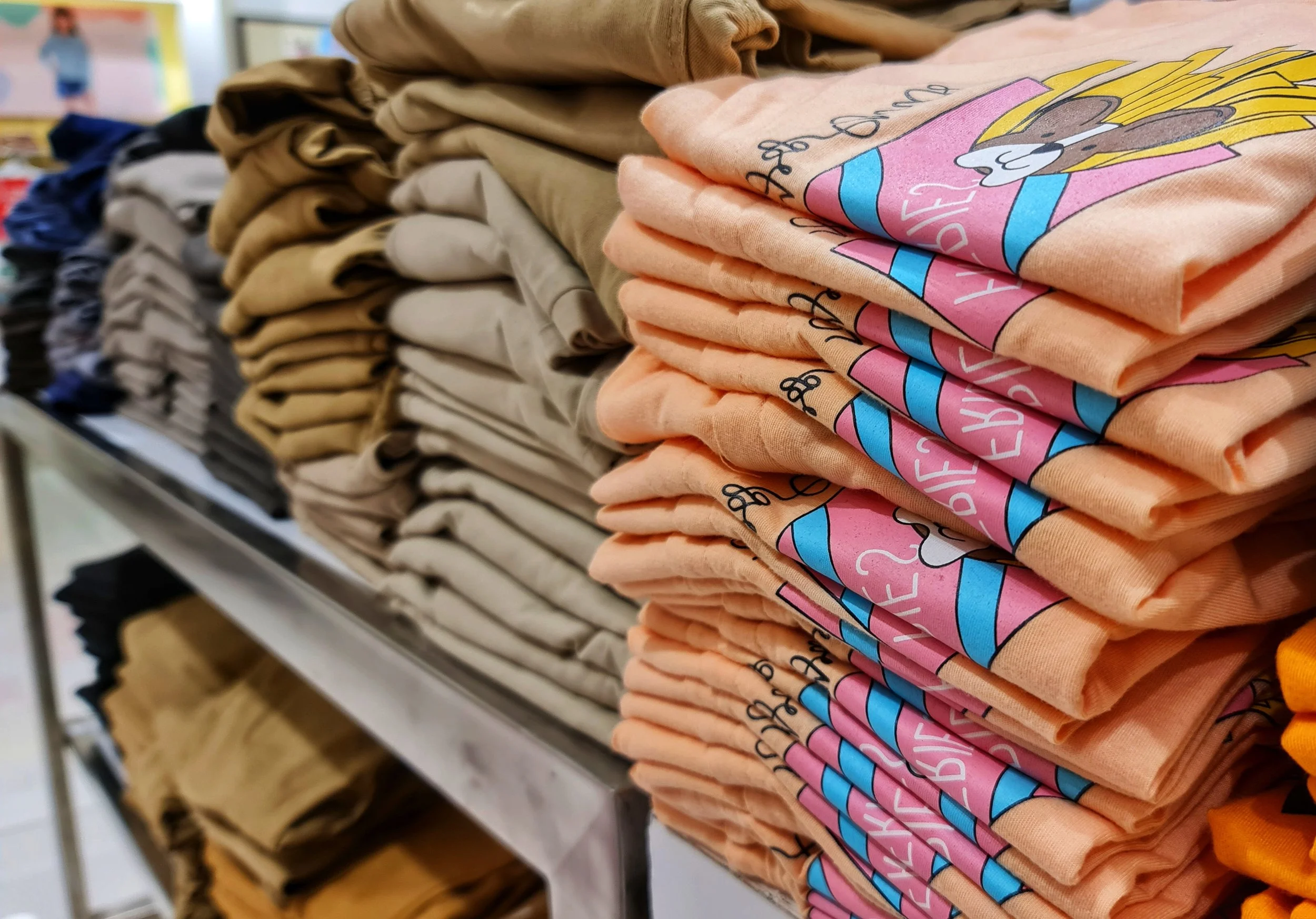 Stacks of folded T-shirts on display, with a pink shirt featuring a cartoon character and colorful design prominently in focus.