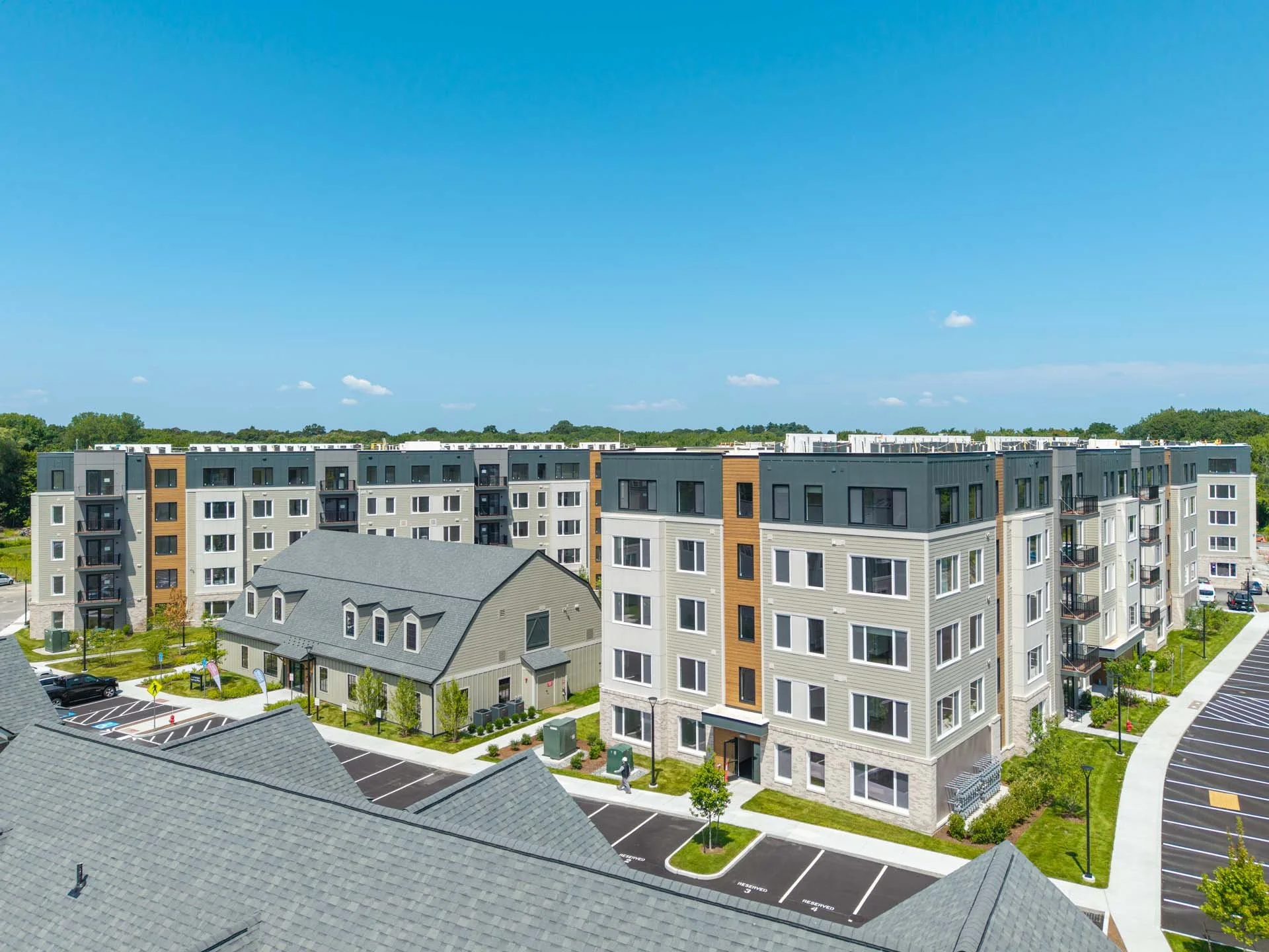 Aerial view of a modern apartment complex with neatly landscaped greenery, parking lot with marked spaces, and a blue sky above.