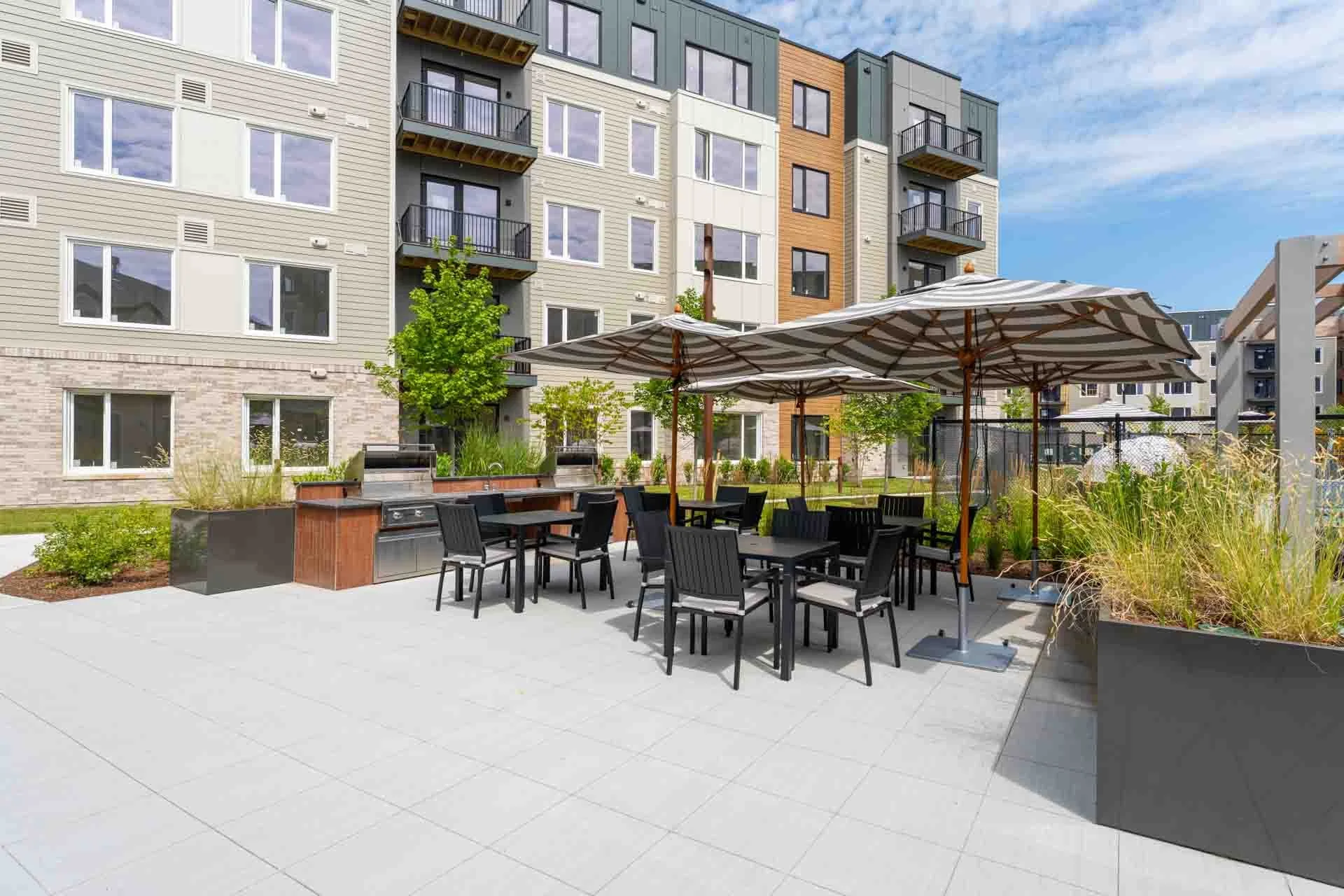 Outdoor communal space with tables, chairs, and umbrellas, surrounded by apartment buildings and landscaping.