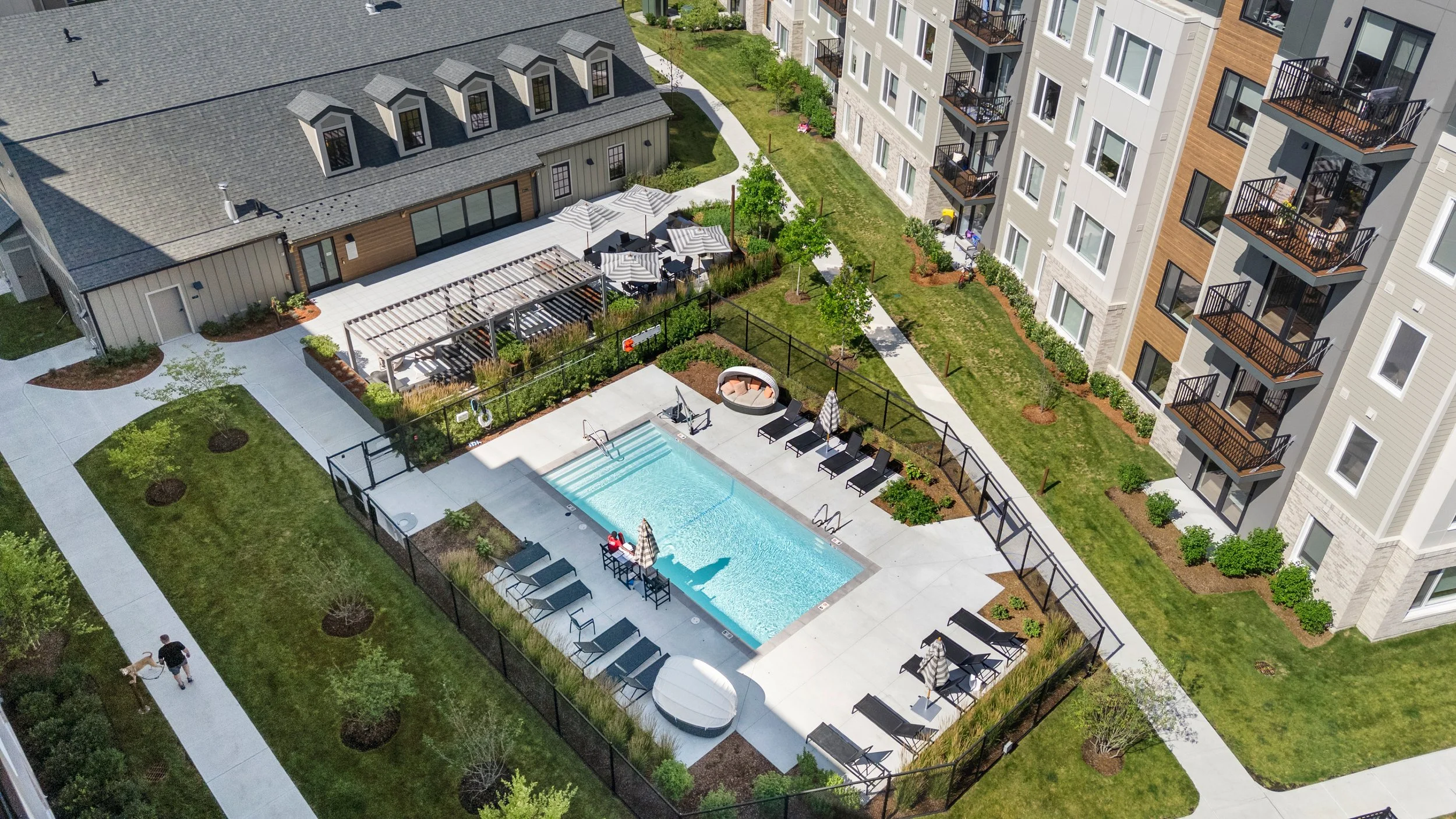 Apartment complex with a swimming pool, lounge chairs, umbrellas, and surrounding greenery under a blue sky.