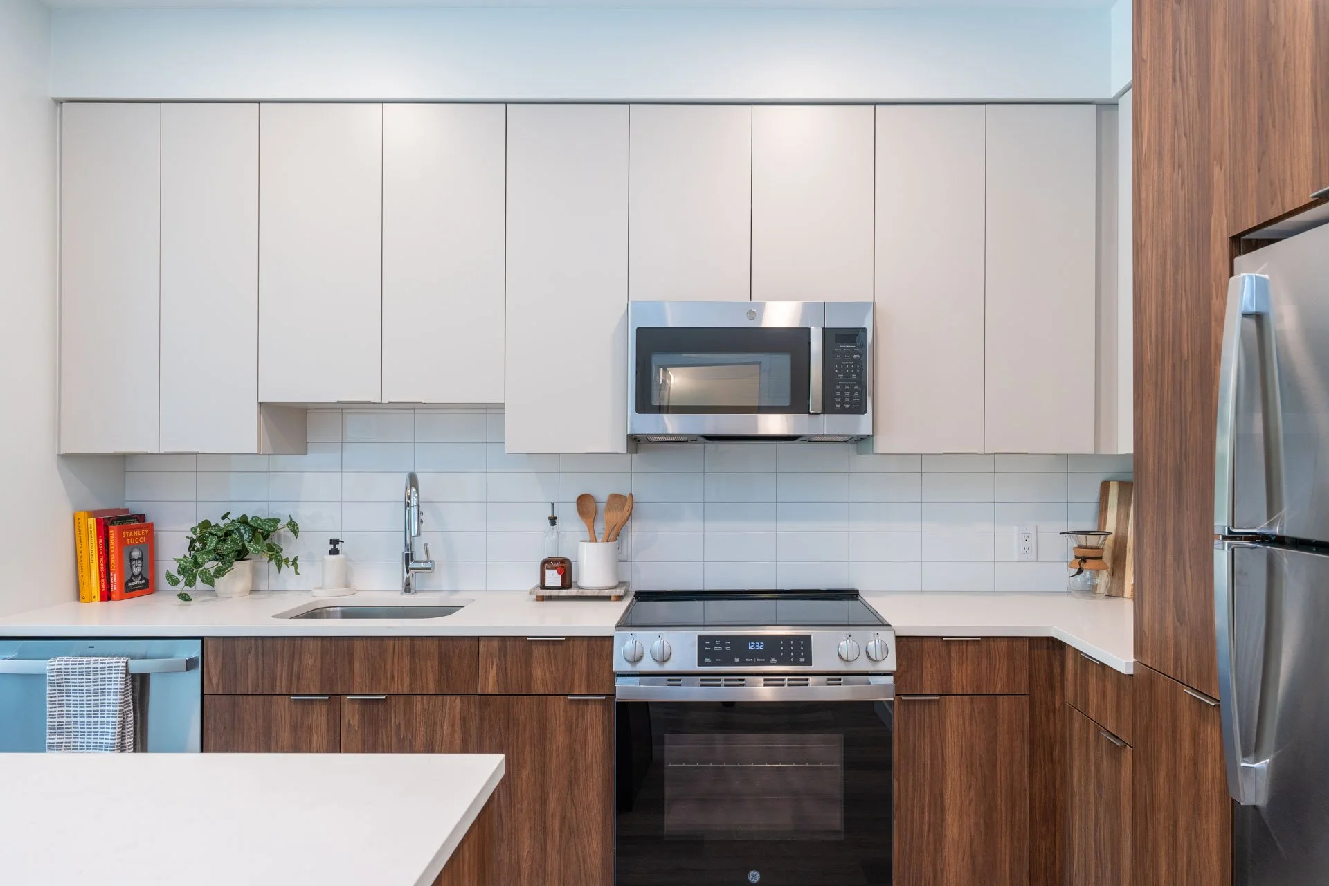 Model apartment modern kitchen with white upper cabinets, wooden lower cabinets, a stainless steel microwave, stove, and refrigerator, white countertop, white tiled backsplash, and decorative items including books, a plant, and kitchen utensils.
