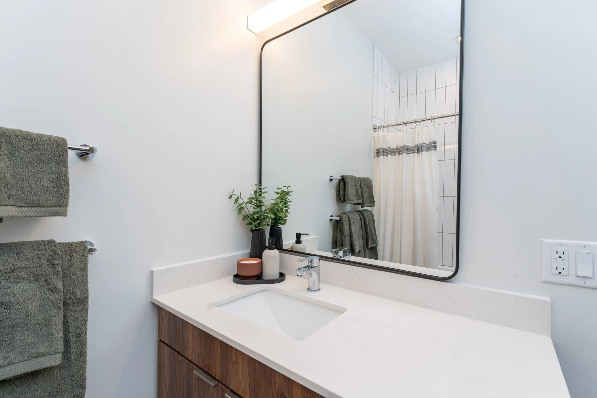 Modern bathroom with a white countertop, a square sink, a large mirror, and a shower with a curtain in the background. There are green towels hanging on a towel rack and a small plant on the counter.