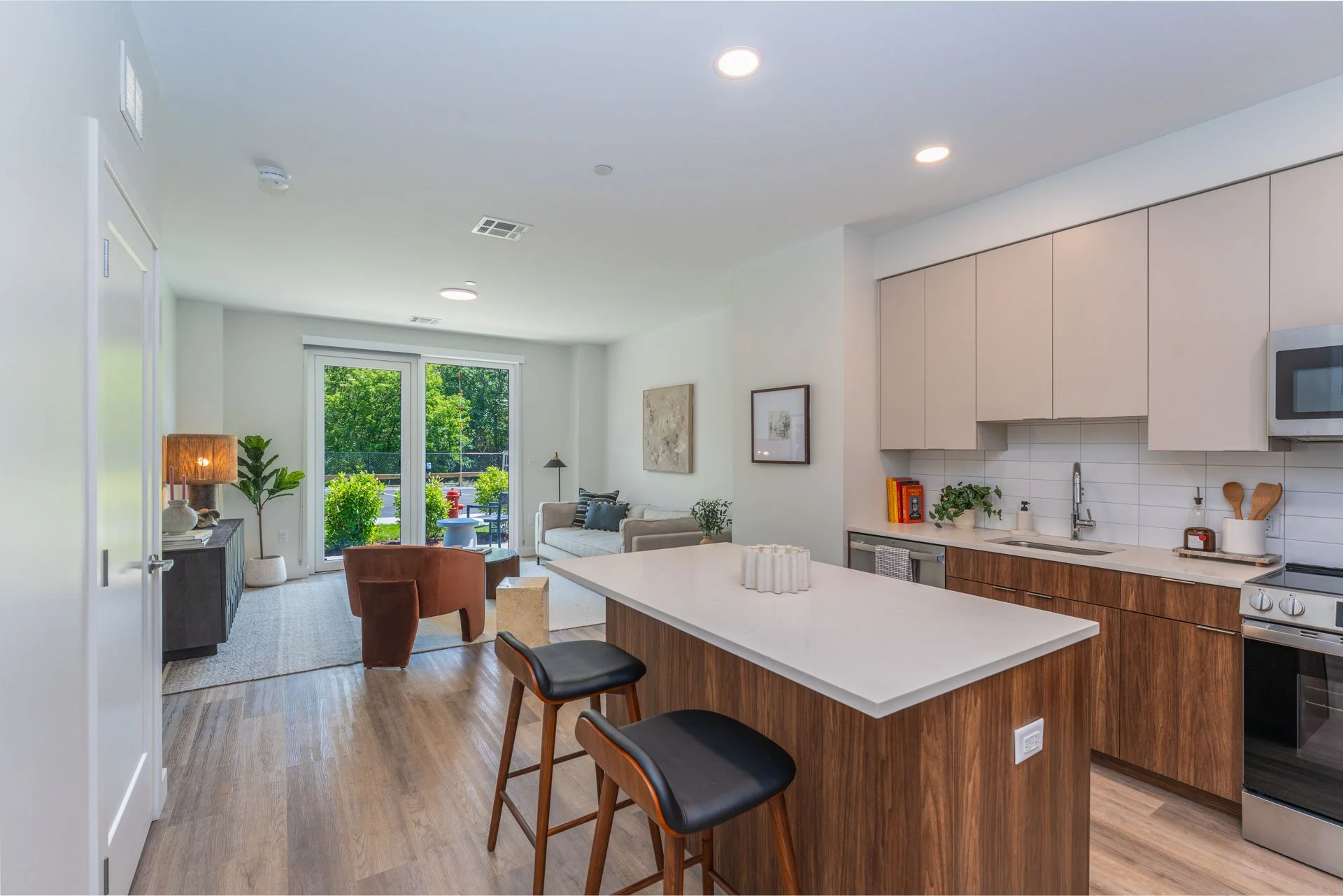 Model apartment open-concept kitchen and living room with sliding glass door leading to a balcony, modern decor, and wooden flooring.