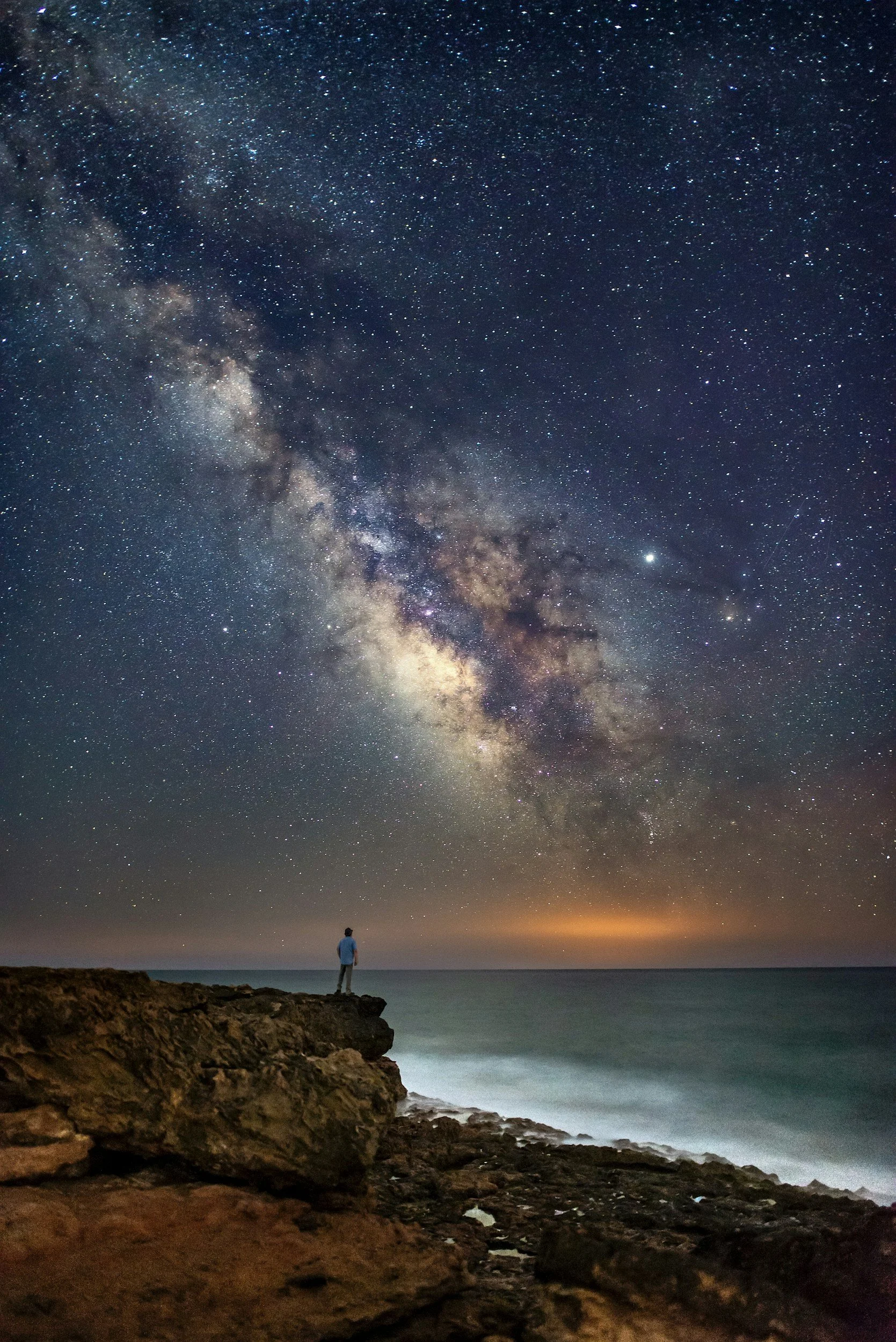man standing on a rock ledge overlooking the ocean underneath the sky with galaxy visible