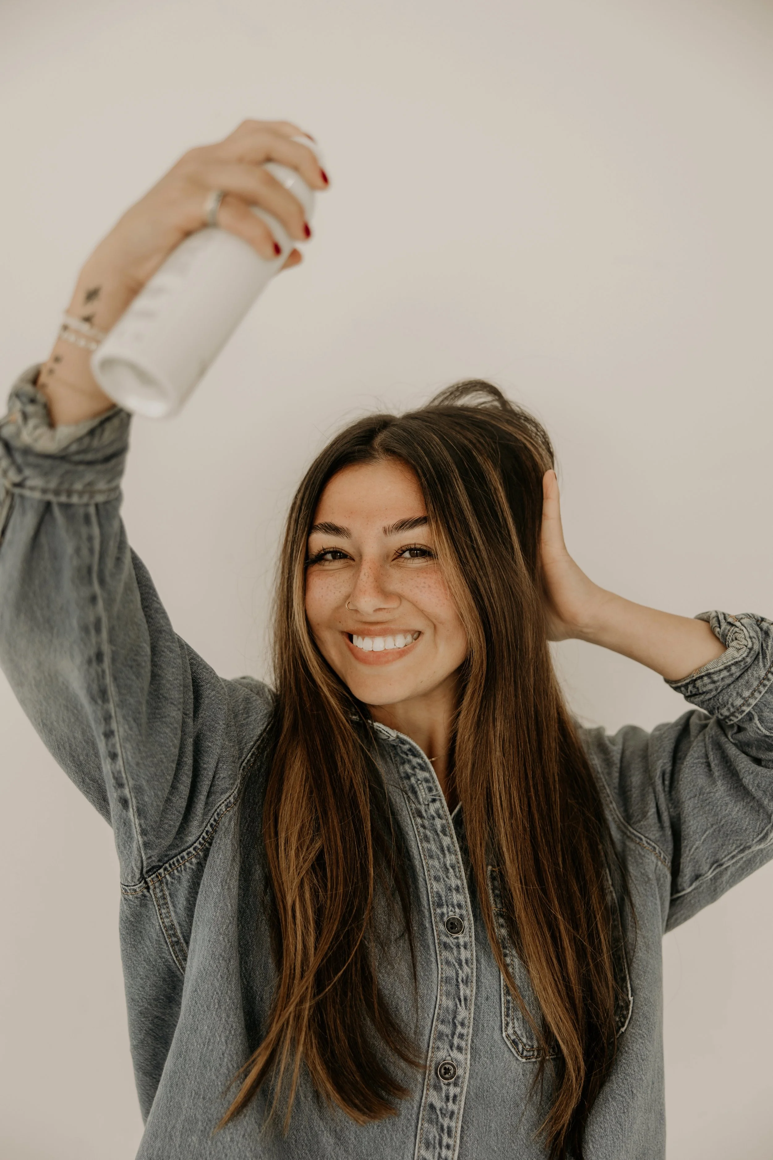A young woman with long brown hair, freckles, and a nose ring smiling and holding a white spray can while posing against a plain light background.