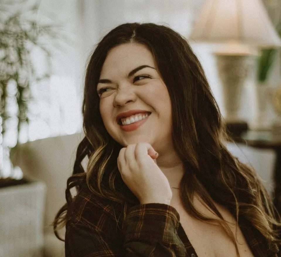 A woman with long dark hair smiling and holding her chin with her hand, sitting indoors with a lamp and a potted plant in the background.
