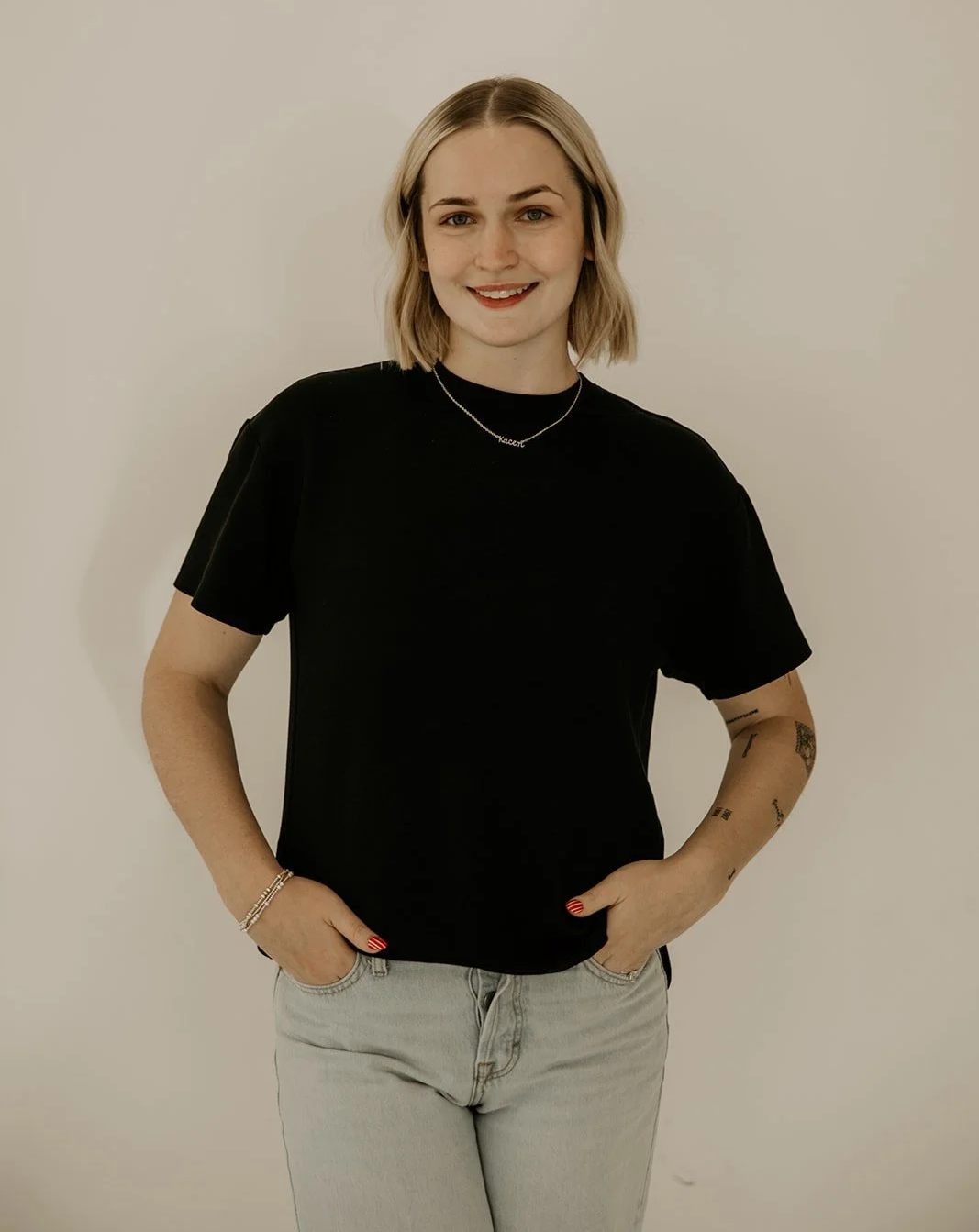 A young woman with shoulder-length blonde hair, wearing a black t-shirt, light-colored jeans, and a necklace, standing against a plain white wall and smiling.