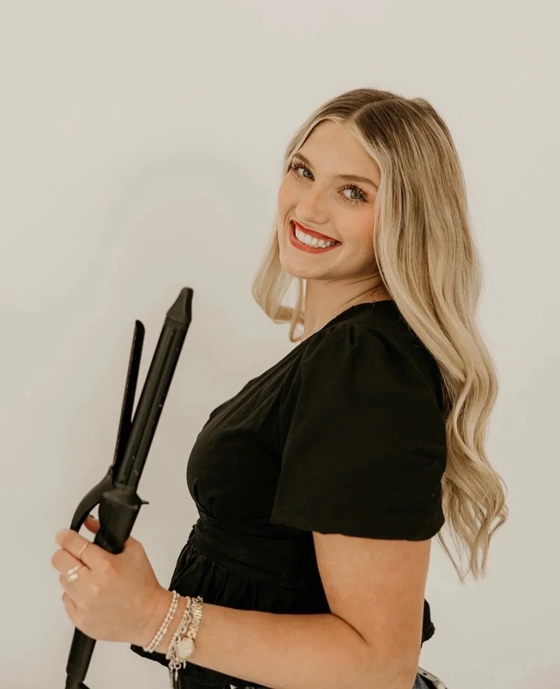 A smiling woman with long blonde hair holding a curling iron in her hand, wearing a black top and bracelets, standing against a plain white background.