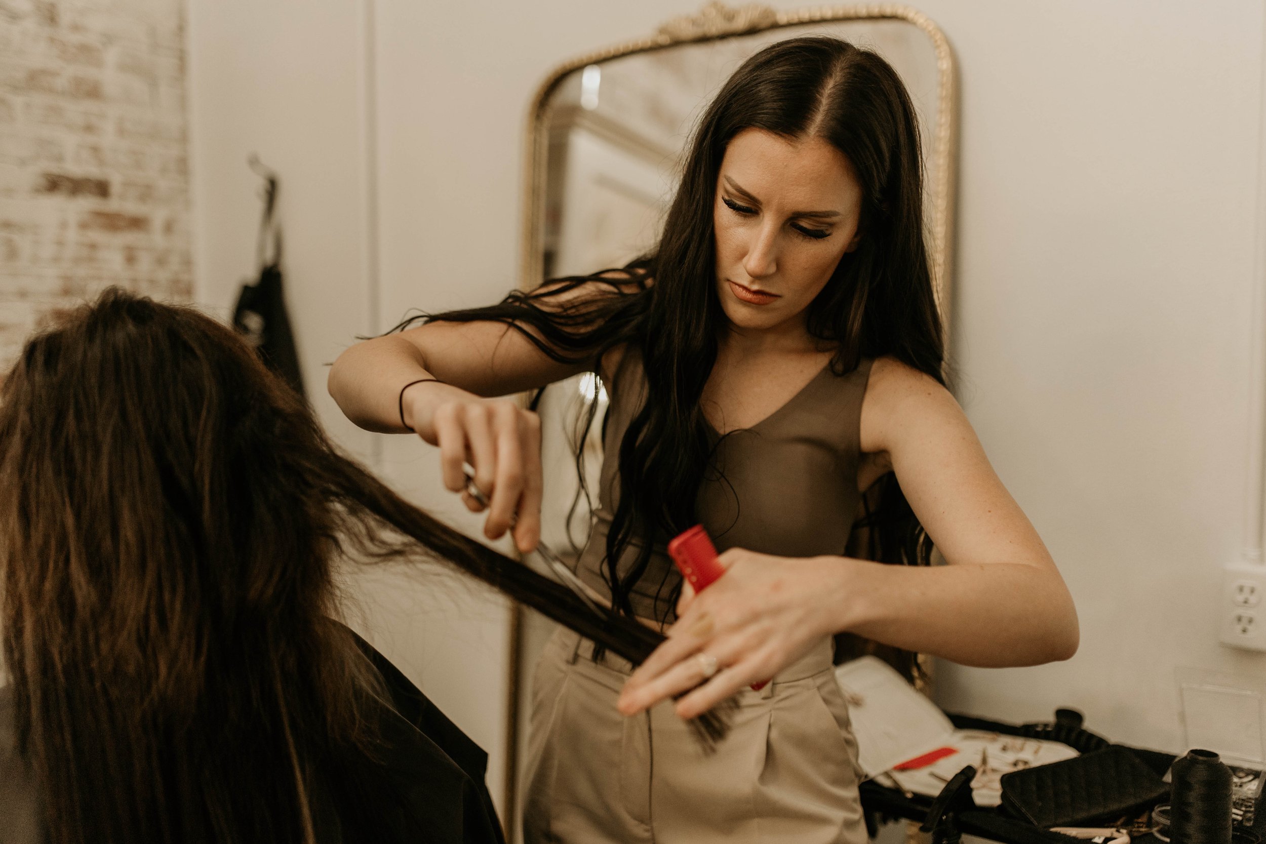 A hairstylist with long dark hair and a sleeveless top styling a client's long brown hair with a hairbrush, in a salon with a mirror and various hair tools on the table.
