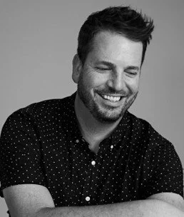 A smiling man with short dark hair, beard, and a patterned short-sleeve shirt, looking down with arms crossed against a plain background.