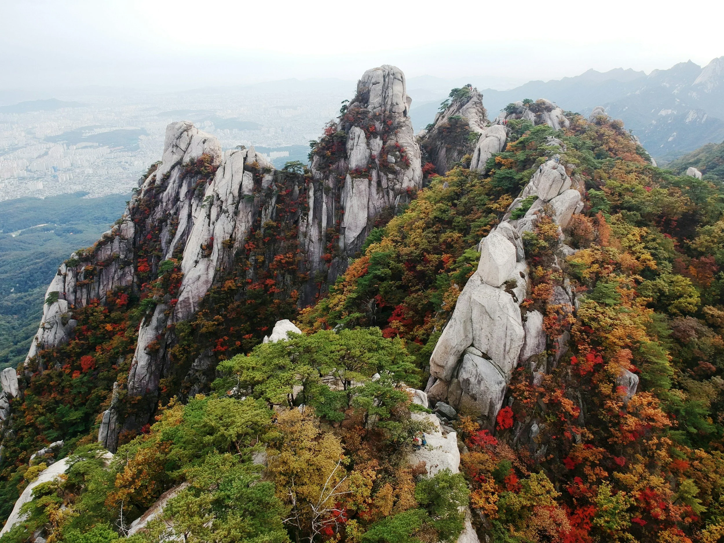 Rocky mountain peak in Korea covered with autumn colored trees overlooking a city in the distance.