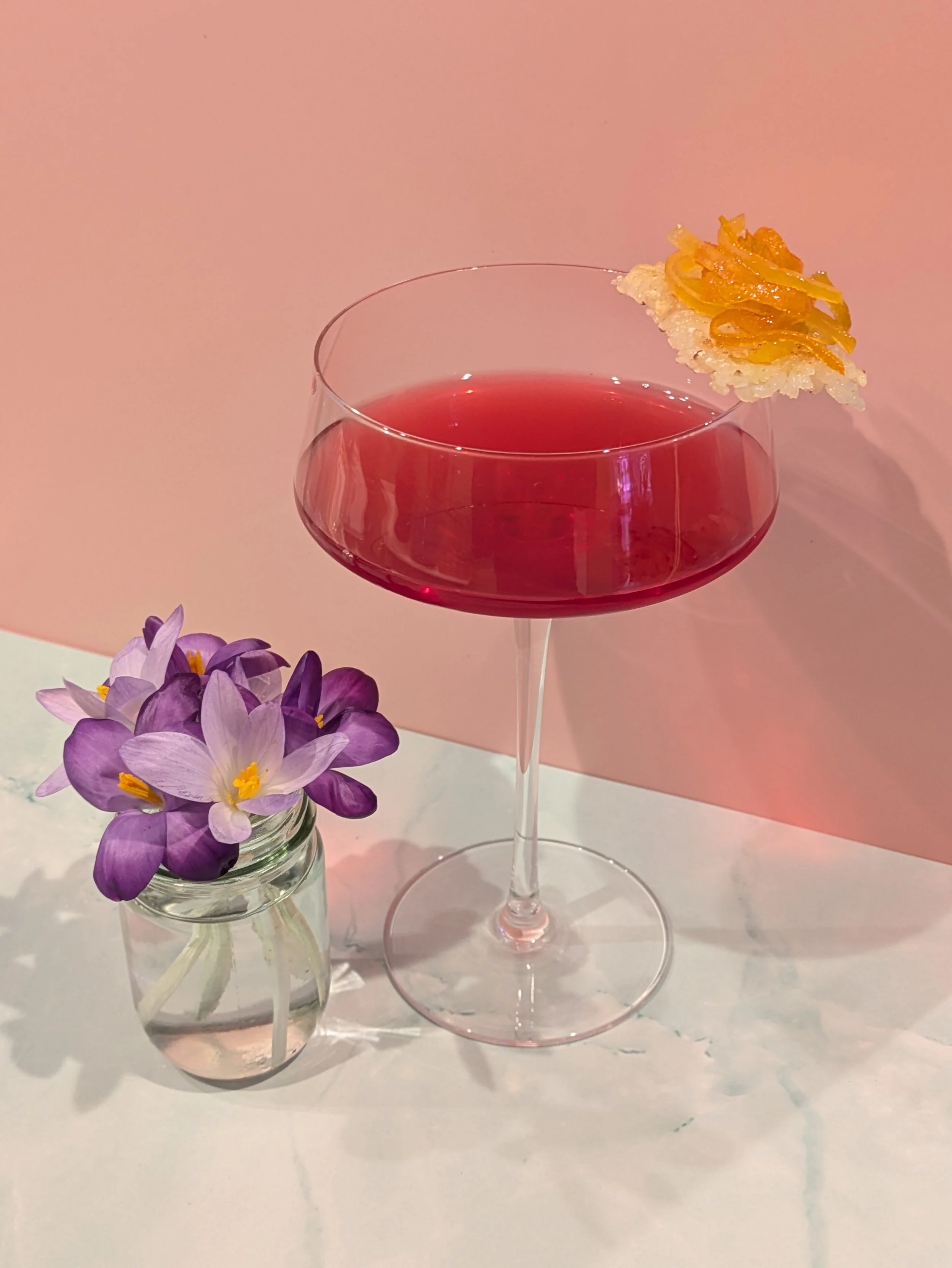 A pink background with a white marble surface holding a pink cocktail in a coupe glass garnished with a crispy rice cracker topped with caramelized yellow fruit or nuts, and a small jar of purple flowers with yellow centers.