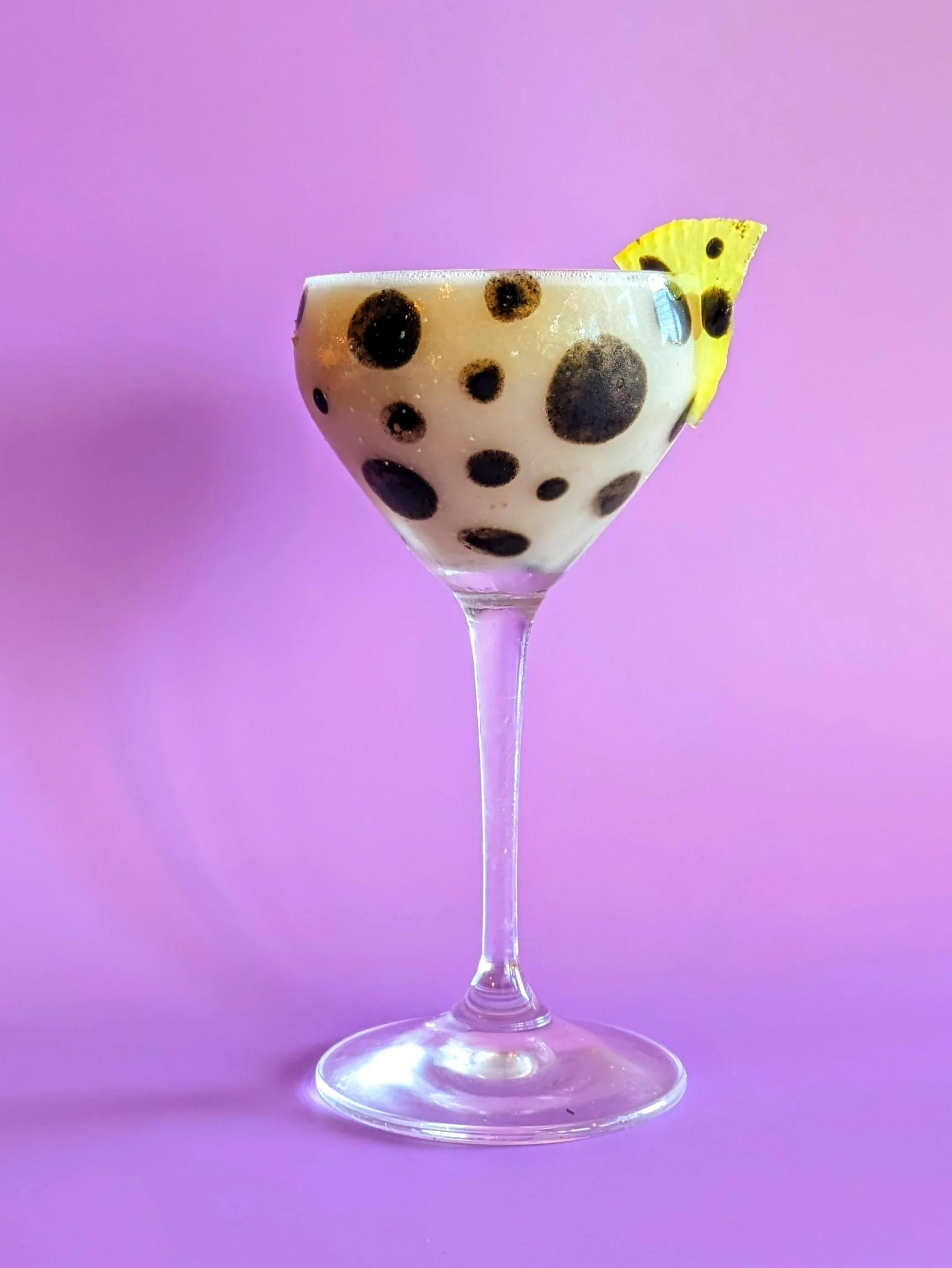 Cocktail in a coupe glass with tapioca pearls and a pineapple wedge garnish against a pink and purple background.