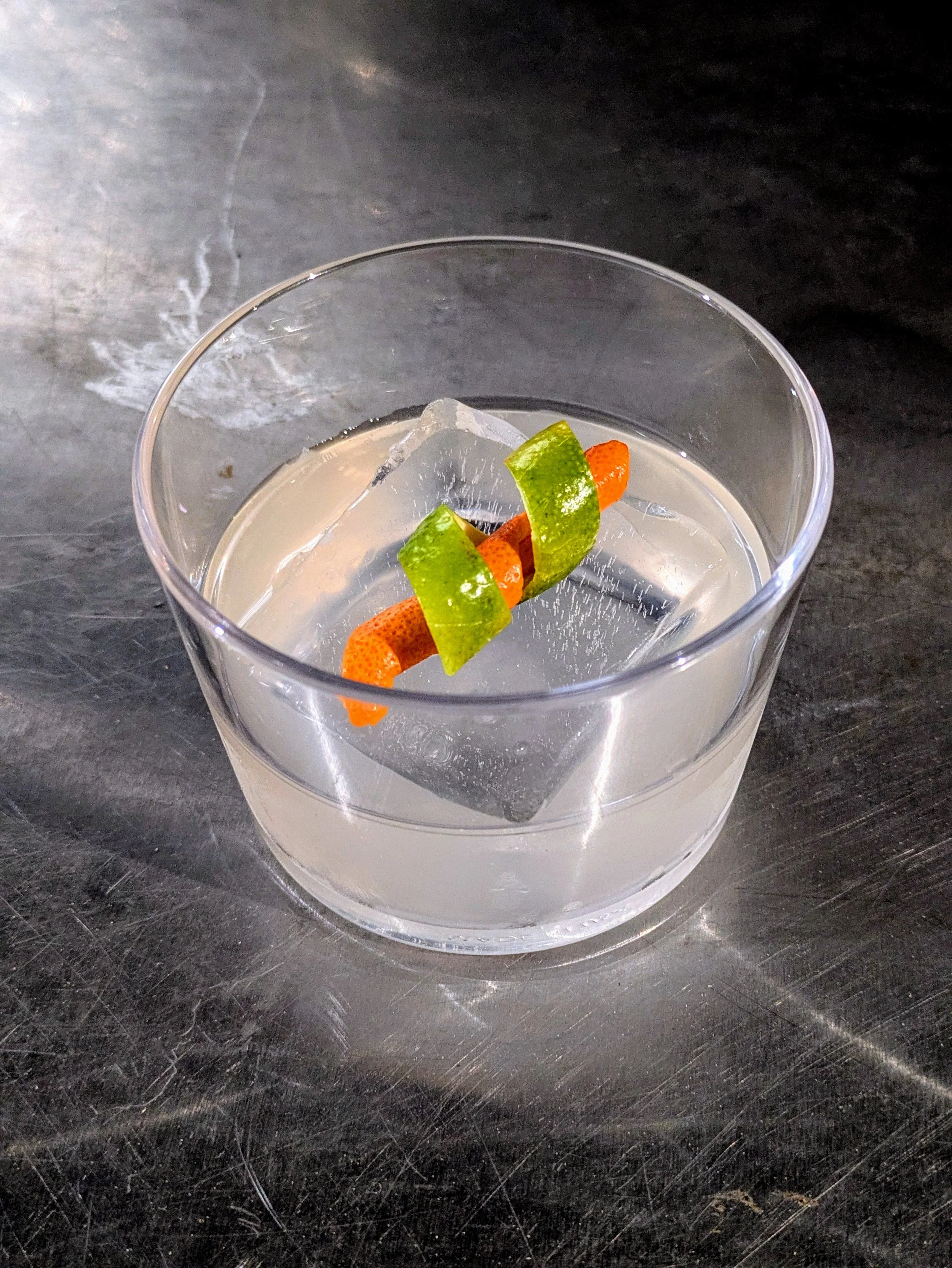 A clear glass tumbler with a large ice cube, containing a clear liquid, garnished with a twist of orange peel and a strip of lemon or lime peel on top. The glass is placed on a dark, textured surface.