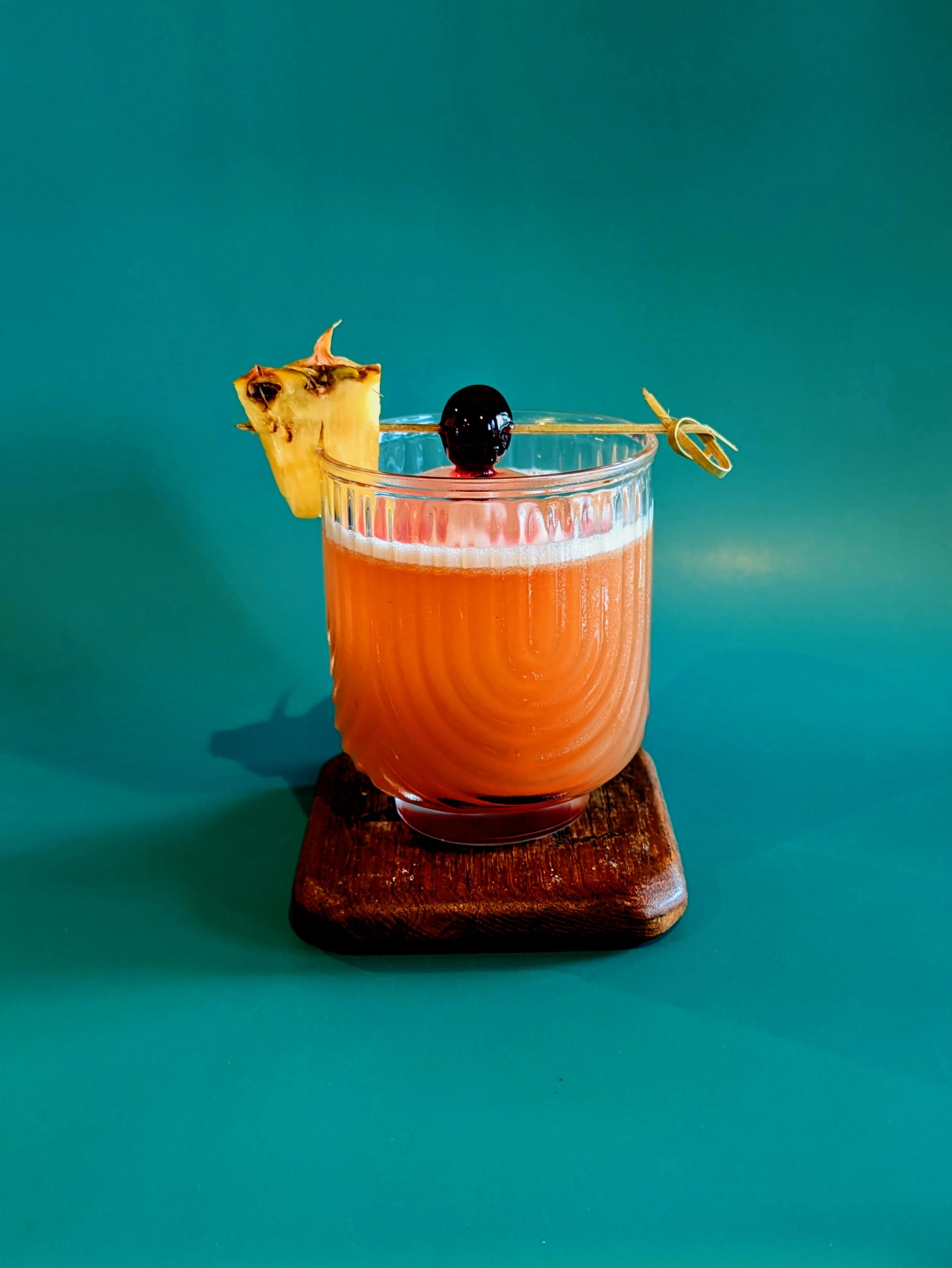 Cocktail with an orange hue in a clear glass, garnished with a pineapple slice and a black cherry on a skewer, set on a wooden coaster against a teal background.
