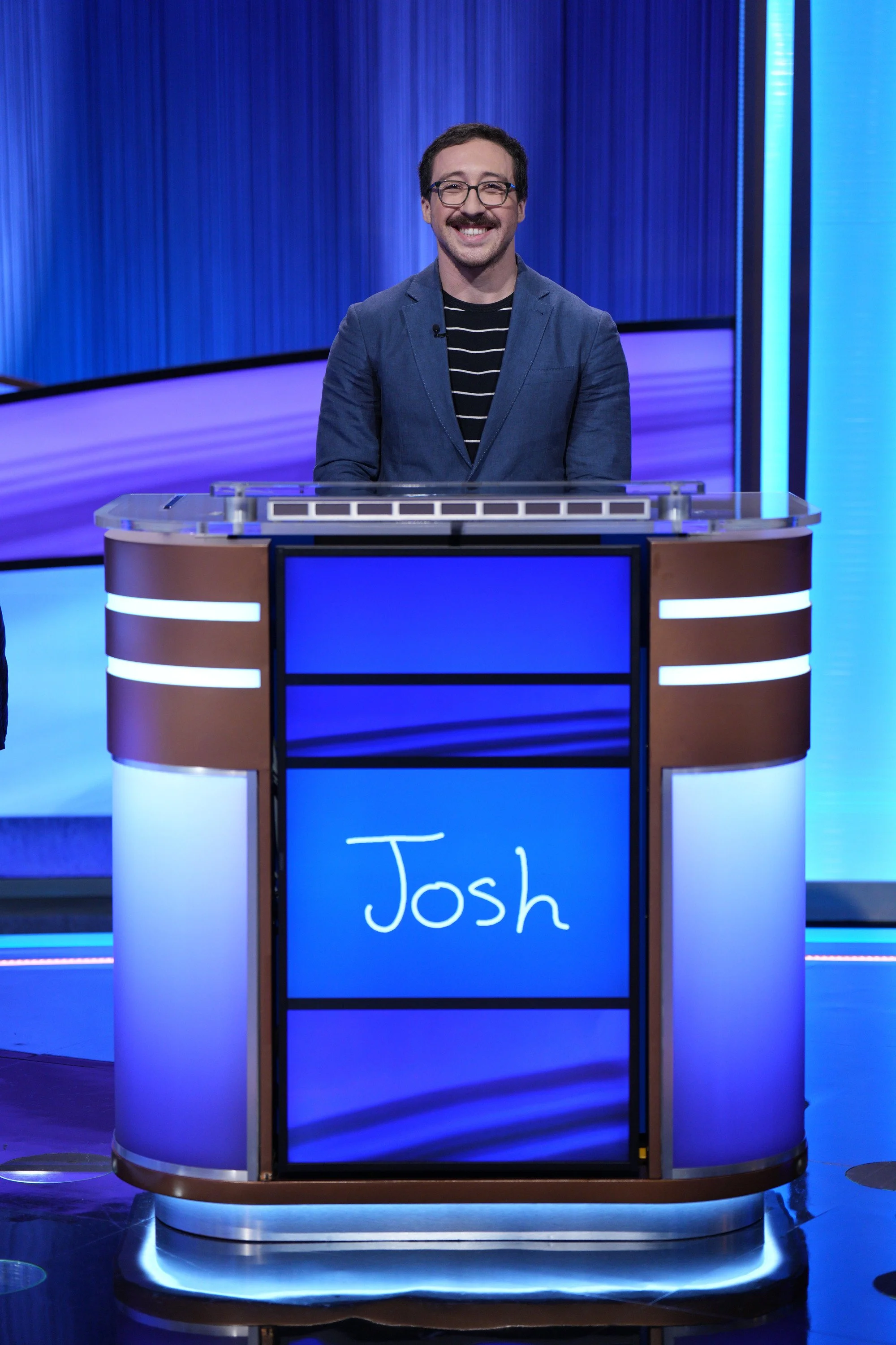 A man with glasses and a mustache wearing a blue blazer and striped shirt standing behind a game show podium with the name 'Josh' on it, on a brightly lit studio set. Jeopardy! game show contestant.