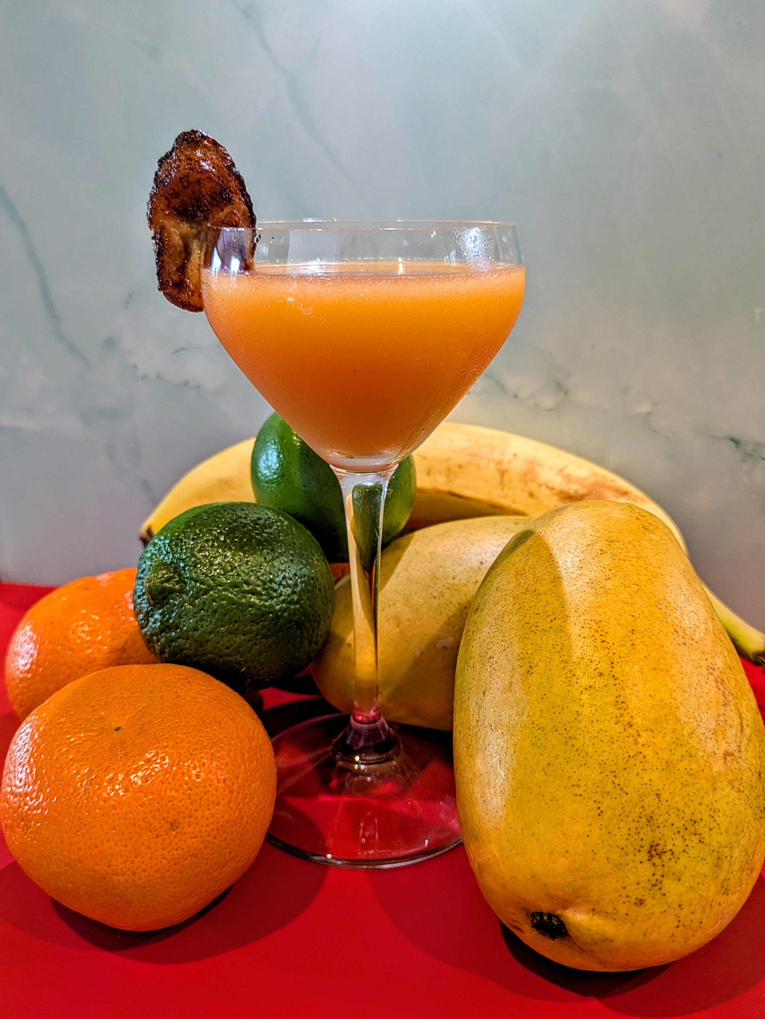 A cocktail in a martini glass garnished with a bacon strip, surrounded by oranges, limes, bananas, and a pear on a red surface against a light wall.