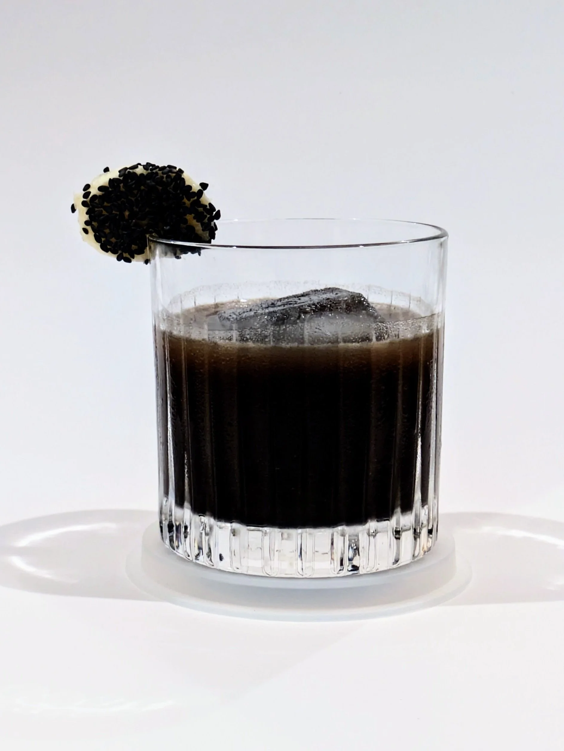 A glass of dark soda with ice cubes, garnished with a black sesame-covered lychee on the rim, on a white surface against a white background.