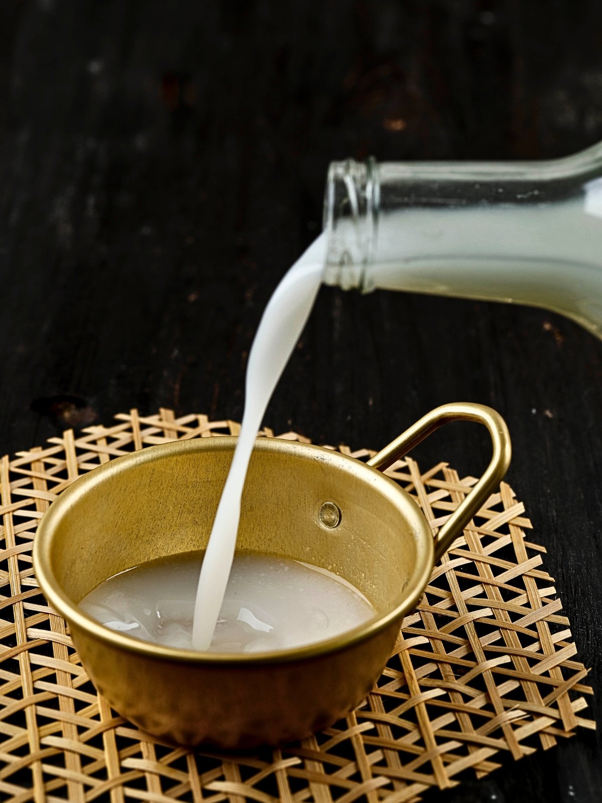 A glass bottle pouring Korean rice wine called makgeolli, a white liquid, into a gold-colored cup on a woven placemat.