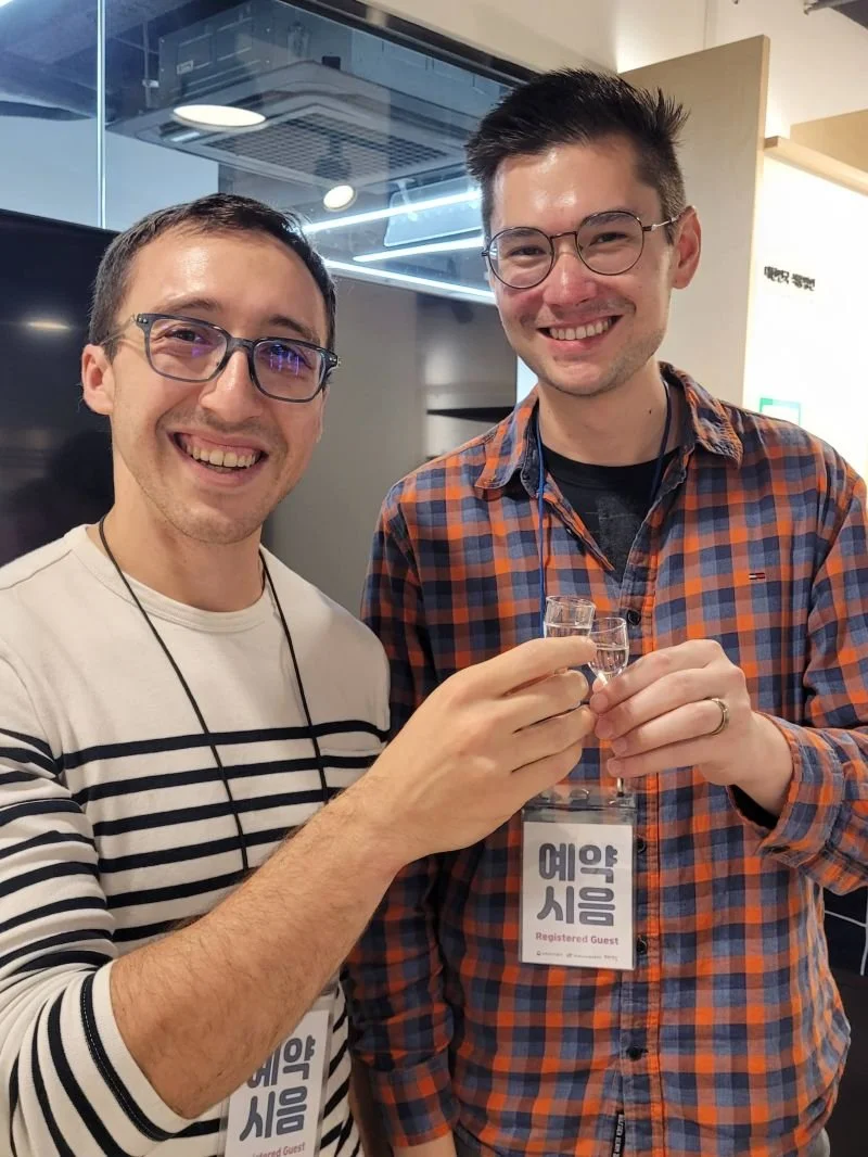 Two men smiling and toasting with small glasses, one wearing a striped shirt and the other in a checkered shirt, at an event with badges that read 'Registered Guest' in Korean.
