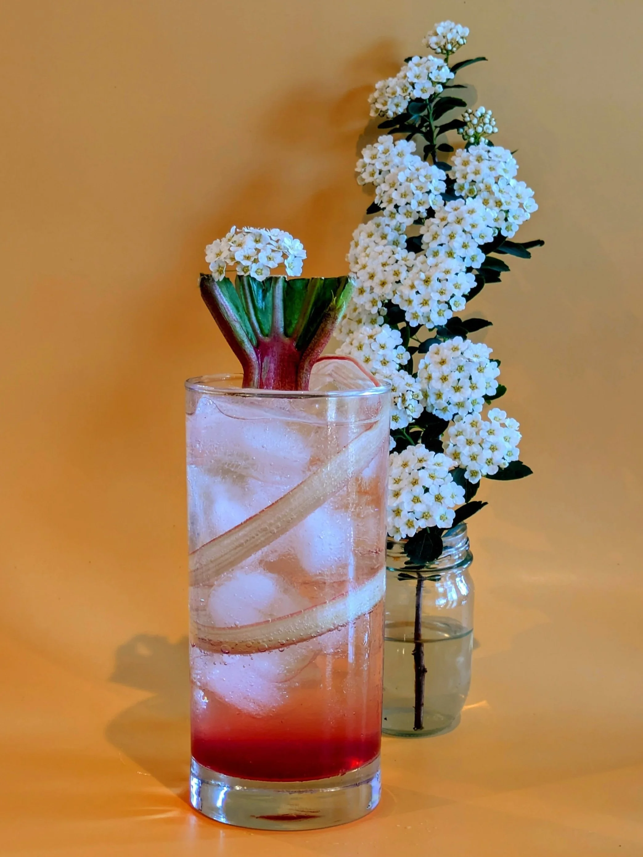 A tall glass with a colorful layered drink, ice cubes, and a slice of fruit, with a star-shaped flower garnish, in front of a vase filled with white flowering branches against a yellow background.