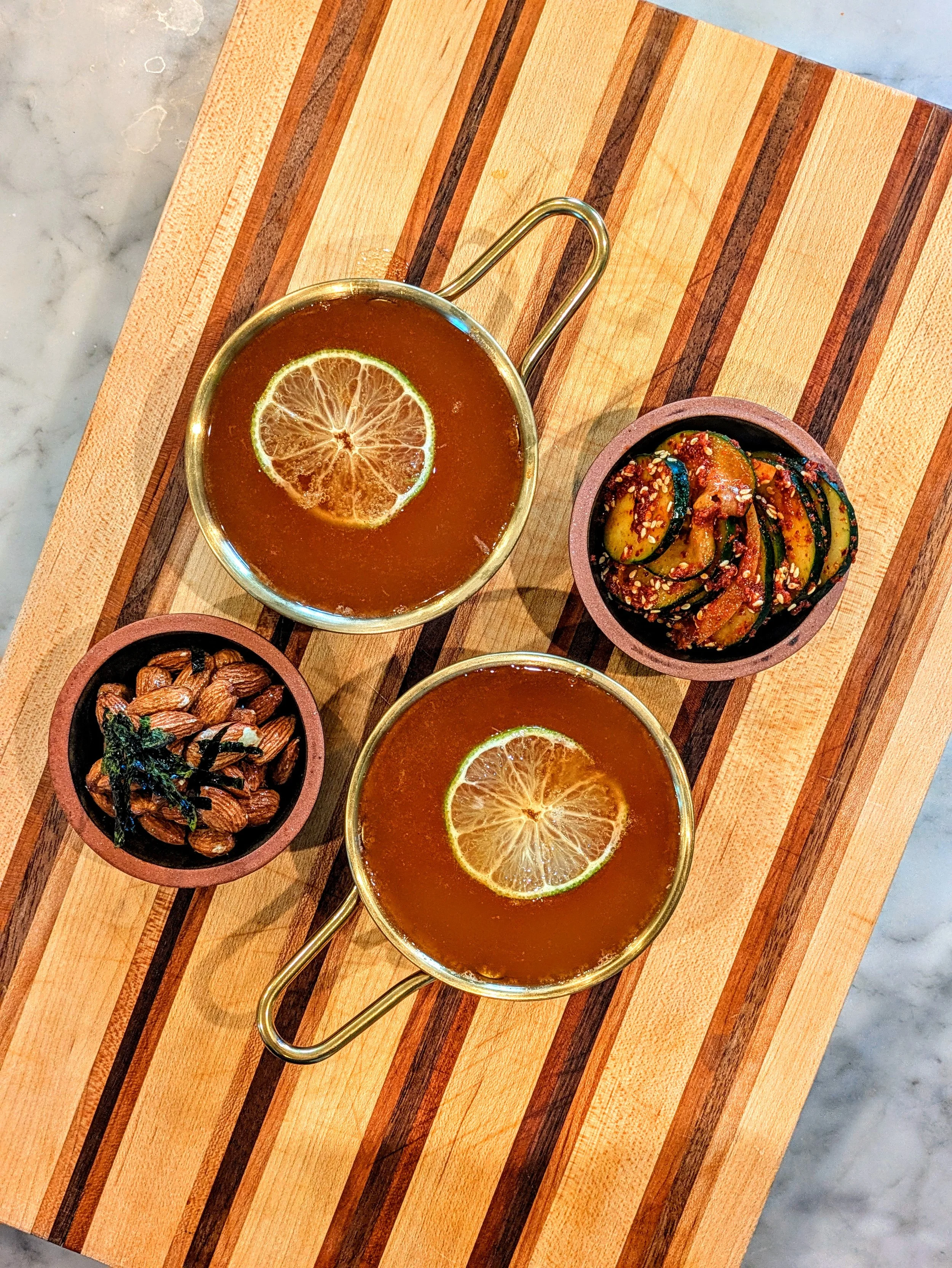 Two glasses of iced tea with lemon slices, two small bowls with cucumber salad and roasted almonds, all on a striped wooden serving board.