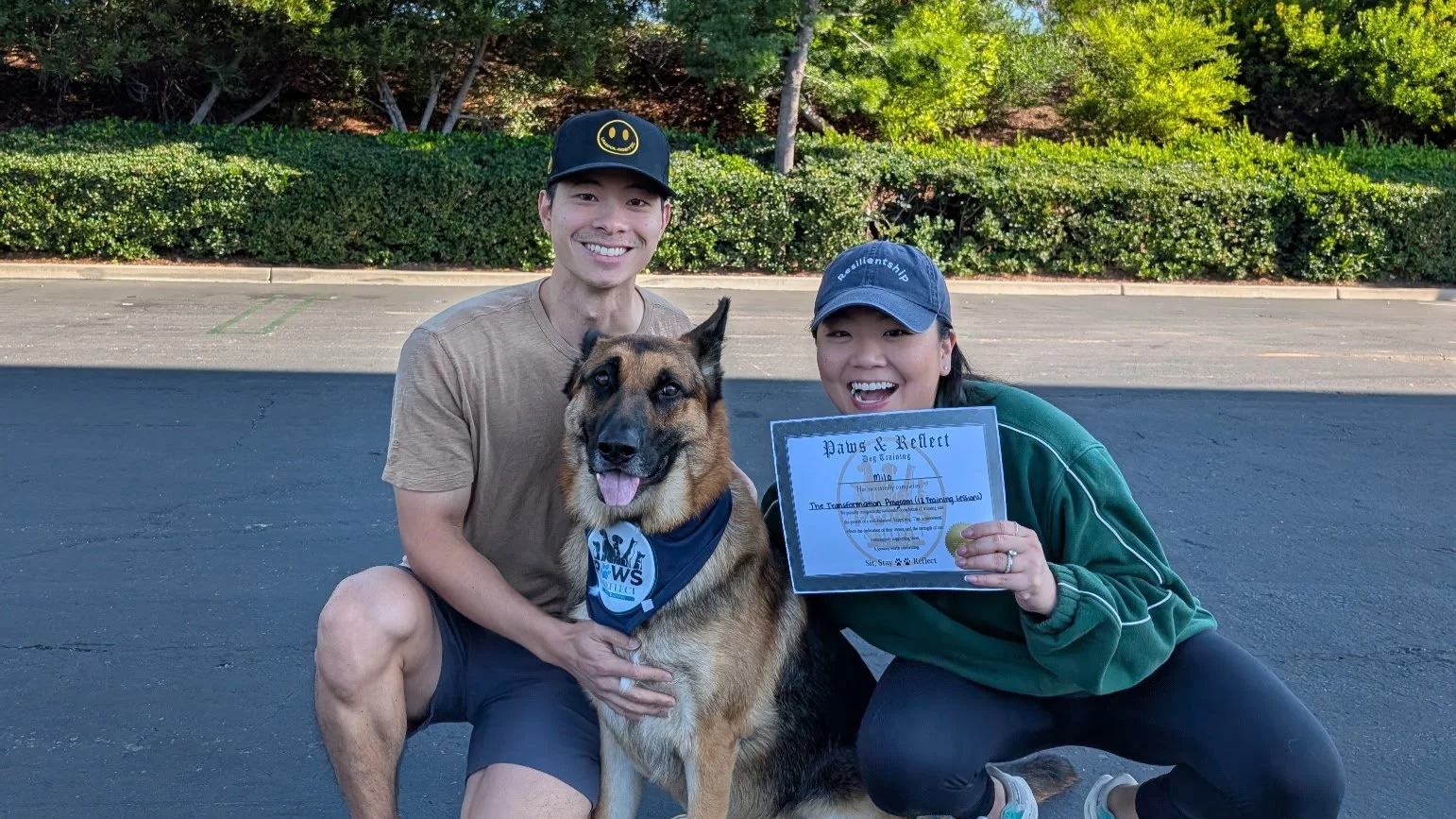 Two smiling people and a dog outdoors in a parking lot, woman holding a certificate, man wearing a black cap with a smiley face, woman wearing a blue cap, greenery in the background.