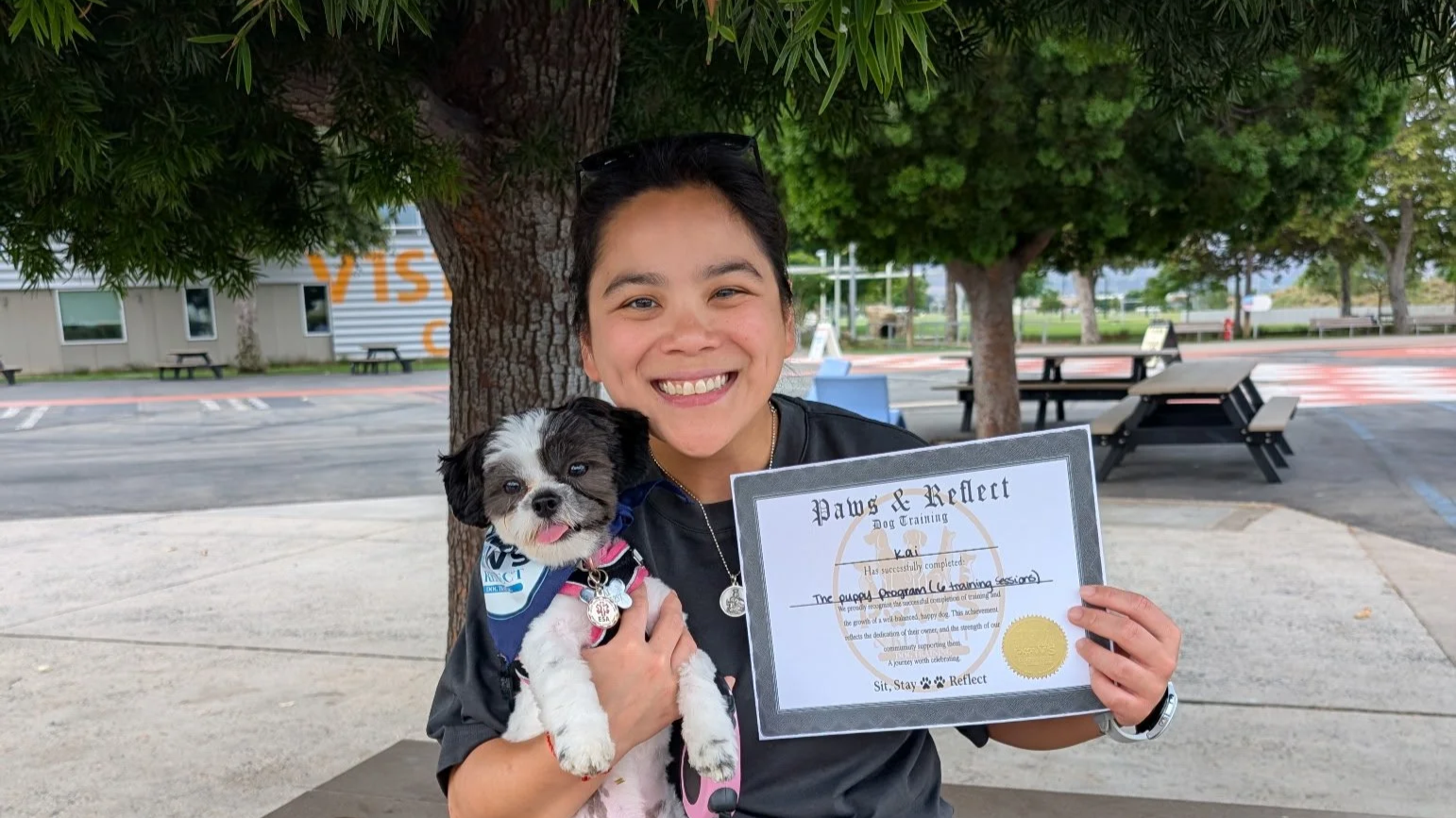 A young woman smiling while holding a puppy and a certificate outdoors under a tree. The woman is wearing a black shirt, and the puppy has black and white fur with a pink collar. There are trees, benches, and a parking lot in the background.