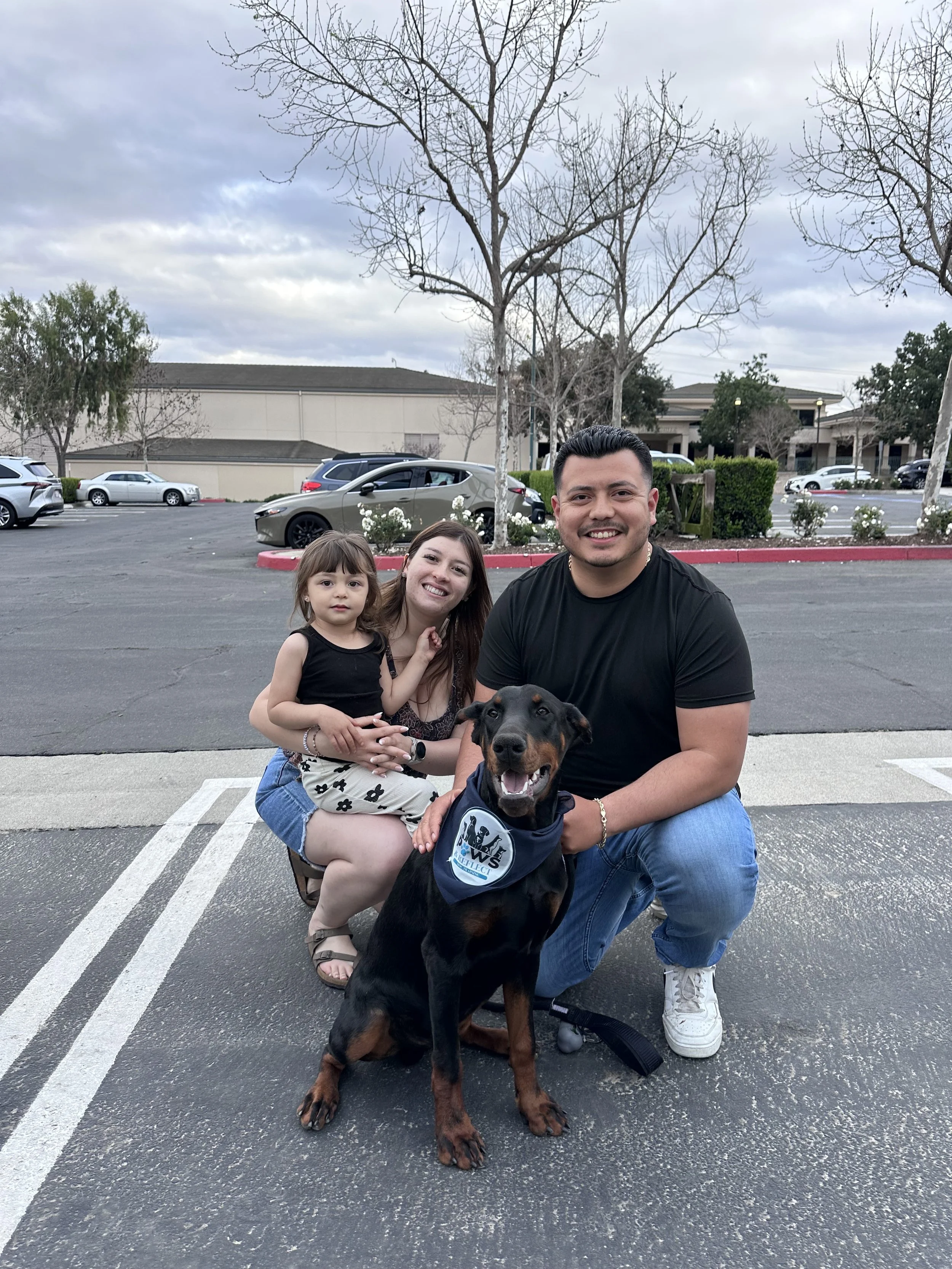 A family of three and a dog posing in a parking lot with cars, trees, and buildings in the background. The family includes a young girl, a woman, and a man. The dog is a Doberman wearing a scarf. It is sitting in front of the man.