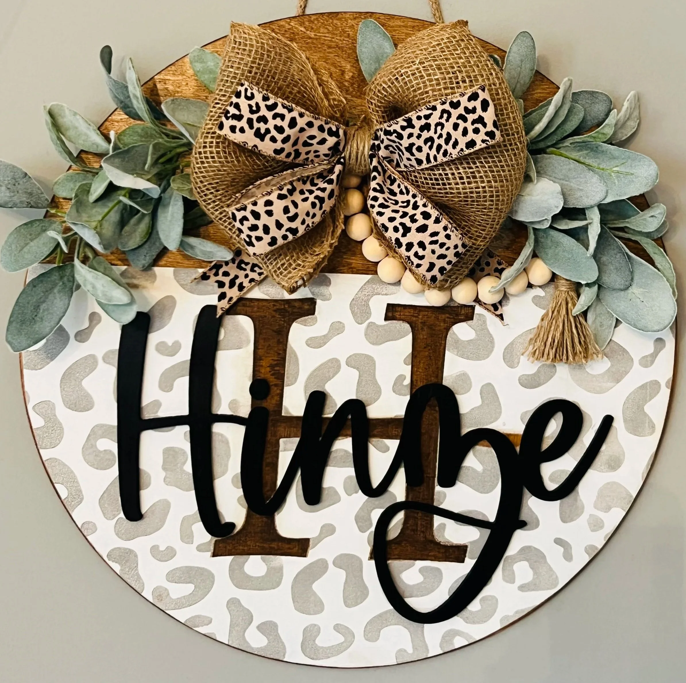 Decorative circular sign with a leopard print bow, eucalyptus leaves, and the words 'Hello' and 'Welcome' in black cursive.