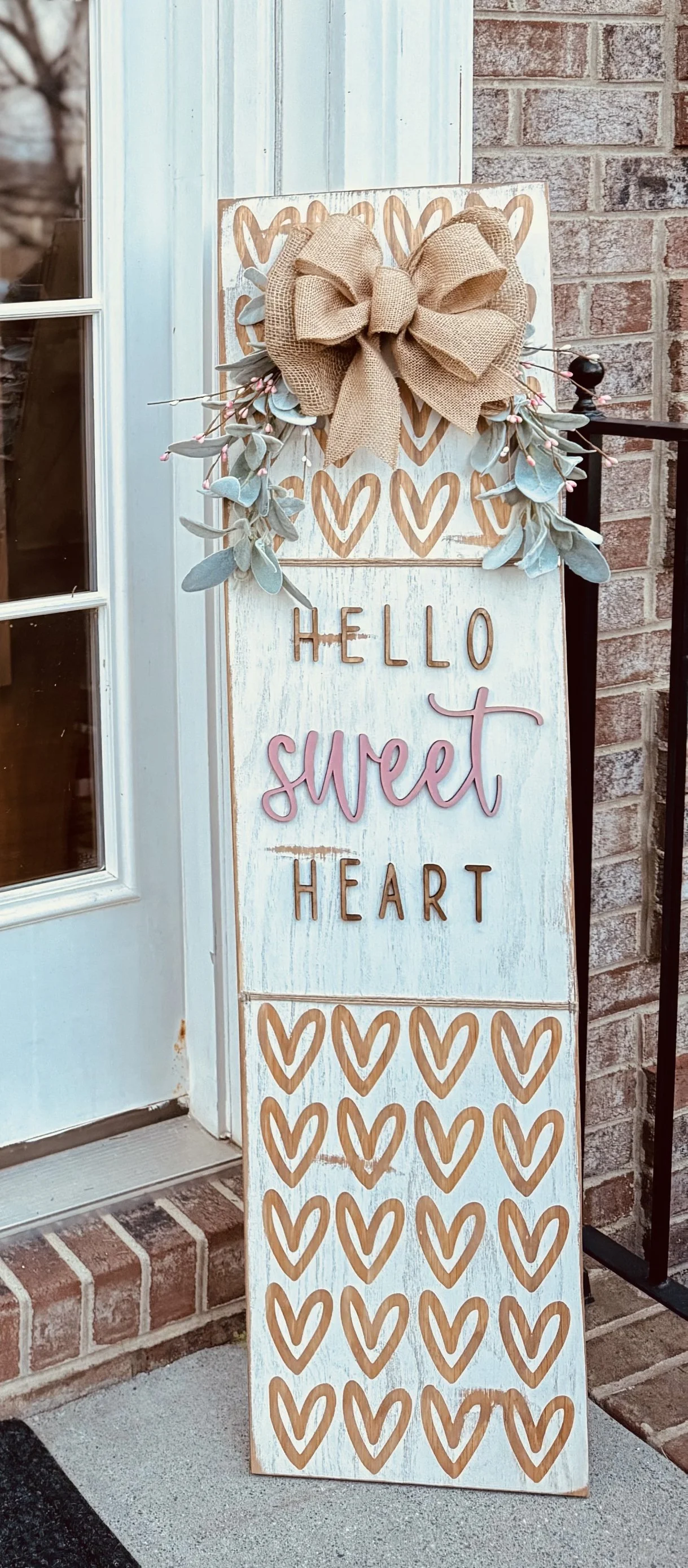 Decorative wooden sign with a large burlap bow and faux eucalyptus leaves, displaying the message 'Hello Sweet Heart' with pink and brown lettering and multiple brown hearts, placed outside near a brick wall and door.