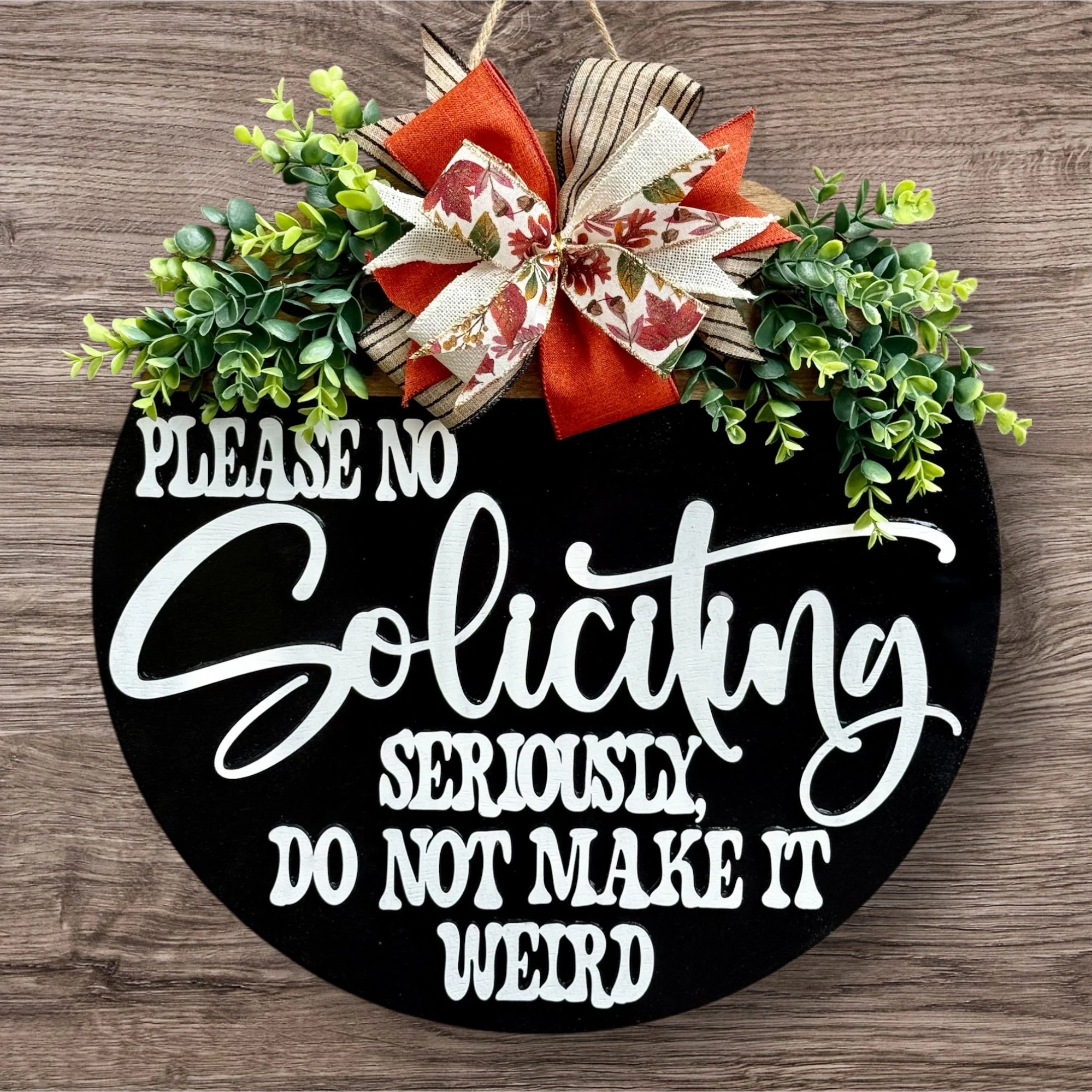Decorative sign with the text 'Please no soliciting seriously, do not make it weird' in white letters on a black circular background, adorned with artificial greenery and a ribbon bow at the top, placed on a wooden surface.