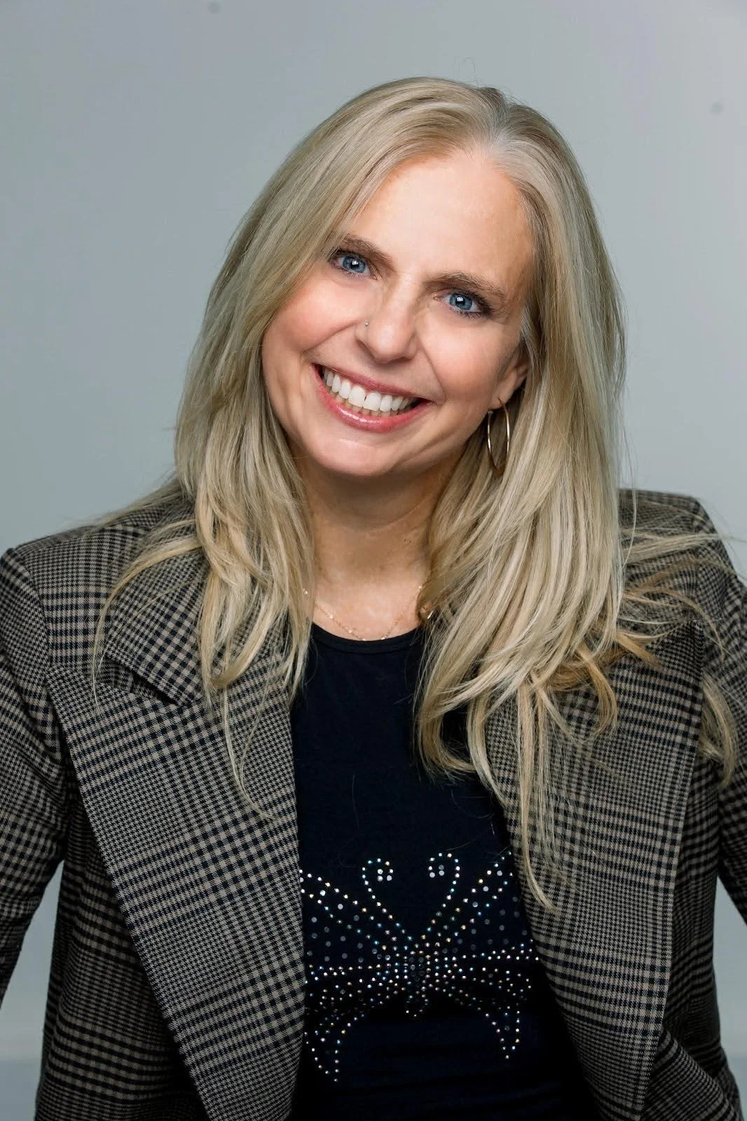 A woman with blonde hair and blue eyes, smiling, wearing a black top with a rhinestone butterfly design and a plaid blazer, against a plain gray background.