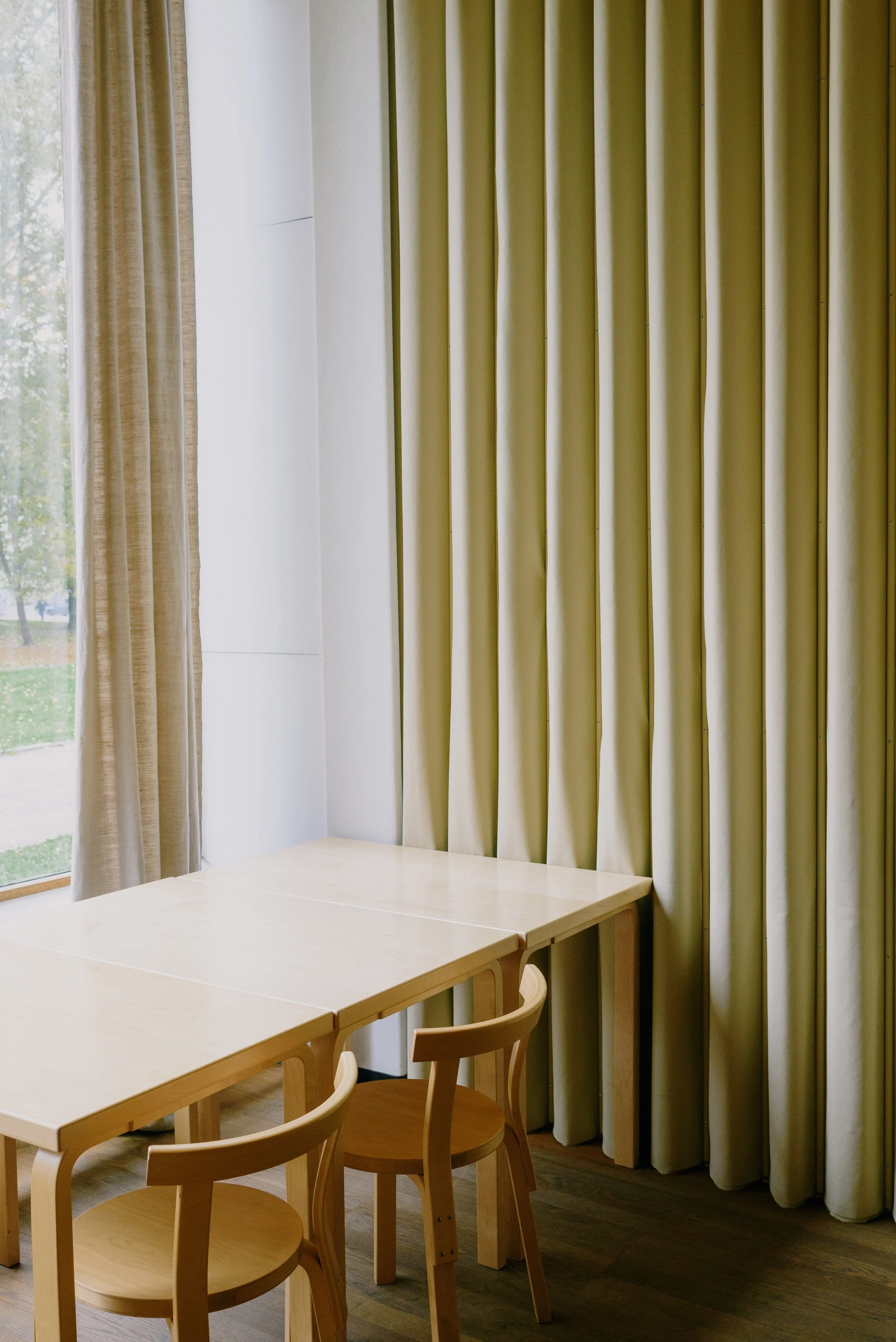 The image shows a room with a wooden table, four wooden chairs, beige curtains, and a window showing a green outdoor scene.