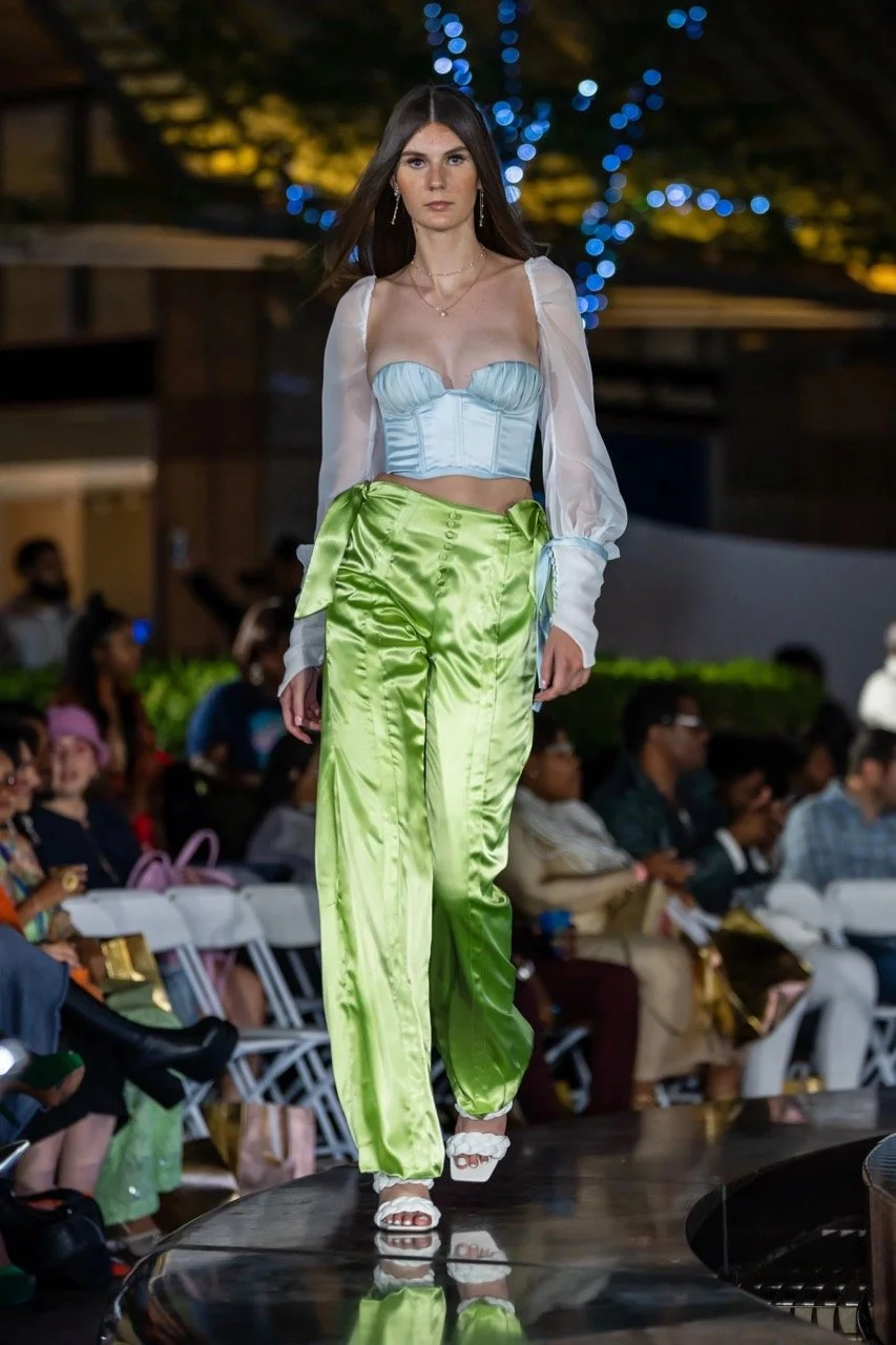 A female model walking down a runway at a fashion show, wearing bright green satin high-waisted pants and a matching strapless top with sheer long mushroom sleeves, with audience members seated on either side of the runway.
