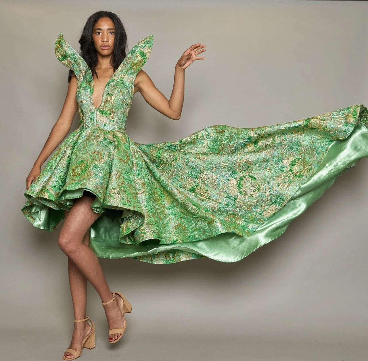A woman in a flowing green and gold gown with high heels, standing against a plain background.