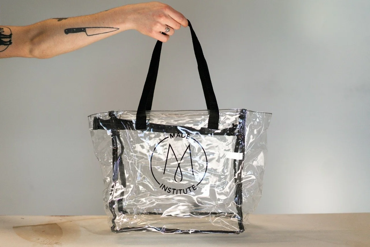 Hand holding a transparent tote bag with black straps and a logo reading 'Made Institute' on the front.