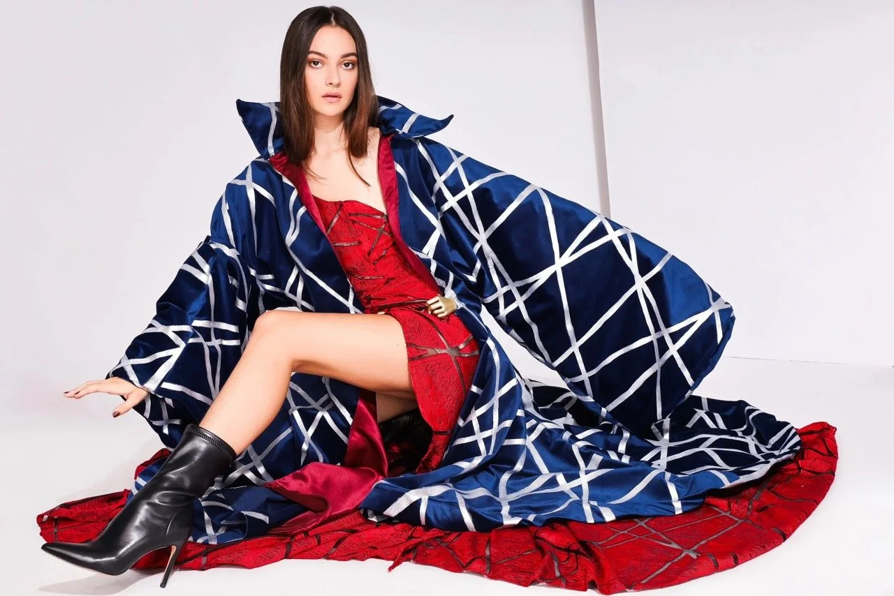 Fashion model sitting on the floor in a red dress, black high-heeled boots, a blue and white checkered robe, with a red quilted blanket beneath her, against a plain white background.