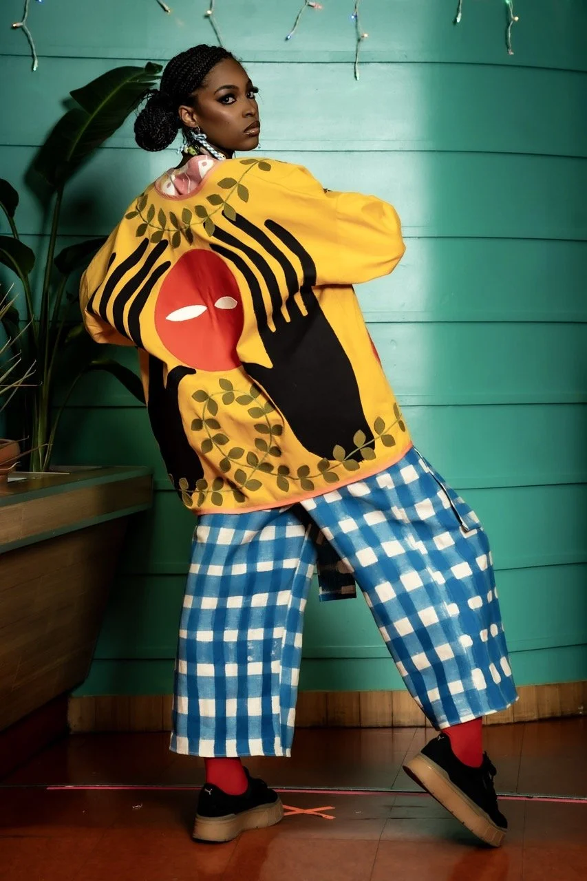 A woman with dark skin and braided hair posing indoors against a turquoise wooden wall, wearing a colorful, abstract printed yellow and black oversized sweatshirt, blue and white checked wide-legged pants, black sneakers, and red socks.