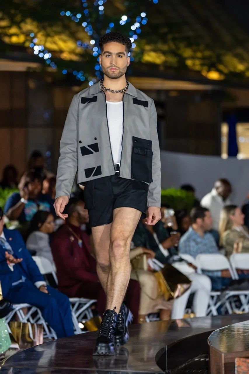 A male model walking on a runway during a fashion show, wearing a gray jacket with black accents, white shirt, black shorts, black combat boots, and a silver chain necklace, with an audience seated in the background.
