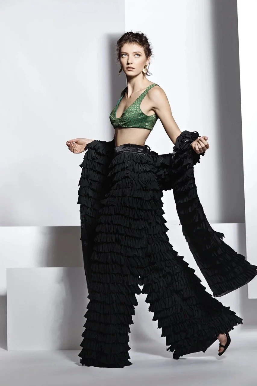 A model wearing a green patterned crop top, wide-leg ruffled black pants, and a matching ruffled black jacket, posing in front of a minimalist white background.